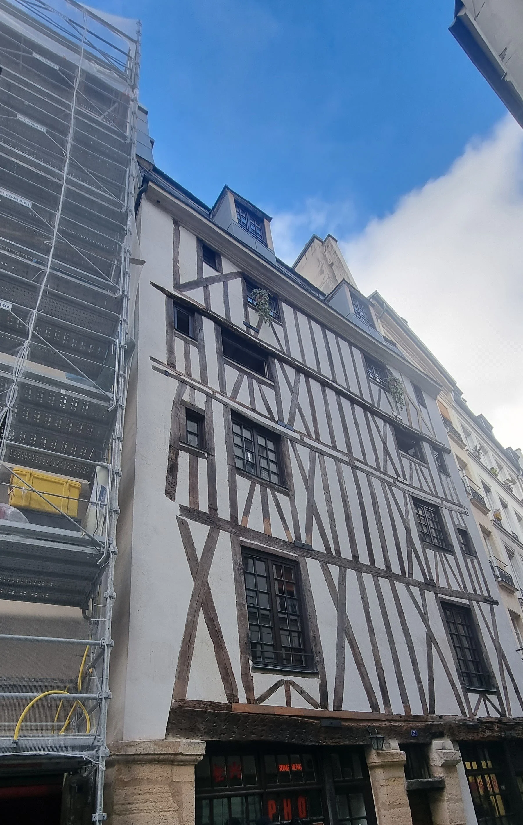 Bâtiment ancien à colombages avec fenêtres à petits carreaux, construction en cours visible à gauche.