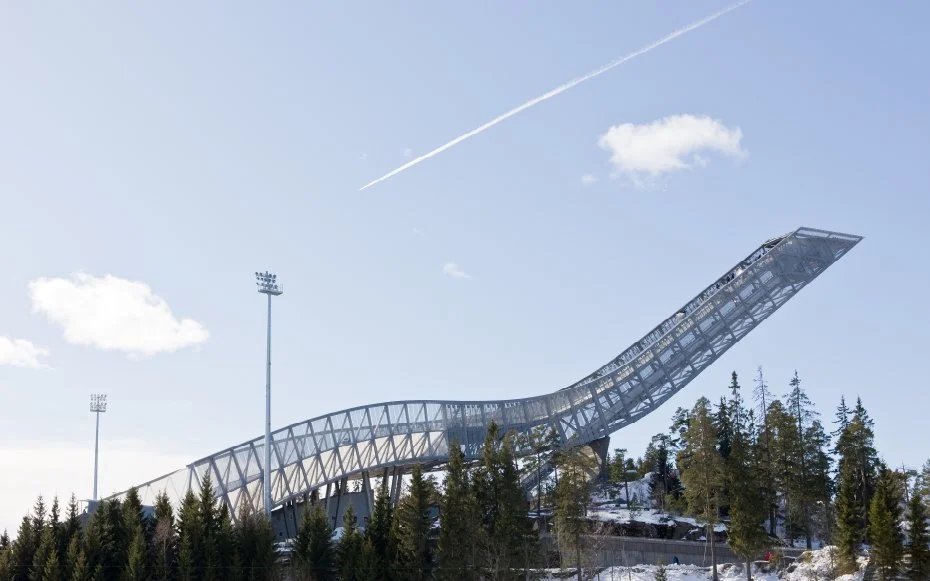 Ski jumping ramp in a snowy area with trees, under a partly cloudy sky with contrail