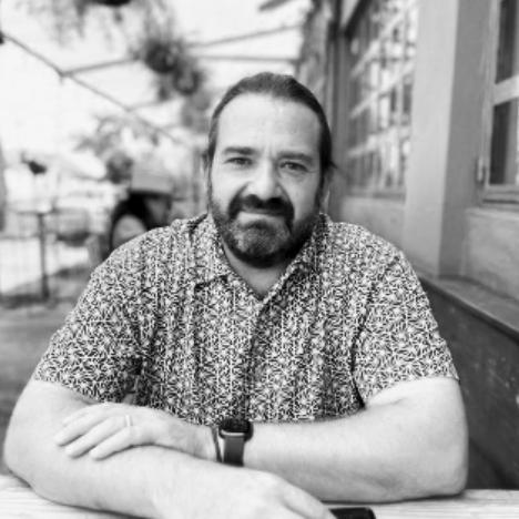 A black and white photo of a man with a beard and long hair tied back, sitting at an outdoor table in an urban setting, wearing a patterned short-sleeve shirt and a watch.