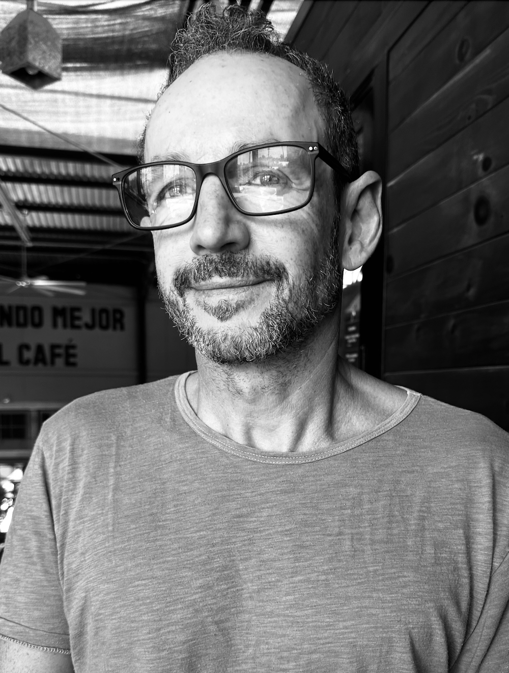 A man with glasses and a beard smiling, sitting in a wooden outdoor area.