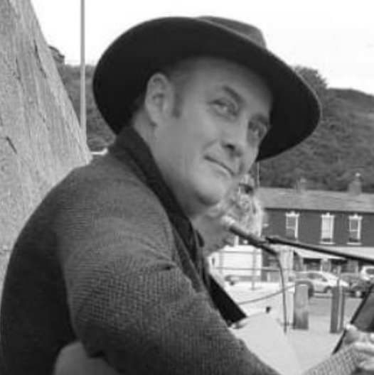 Black and white photo of a man wearing a wide-brimmed hat, sitting outdoors in front of a wooden fence with houses and hills in the background.