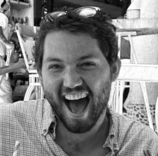 A man with dark hair and a beard, smiling widely with mouth open, wearing a checkered shirt, sitting outdoors at a table with chairs in the background.