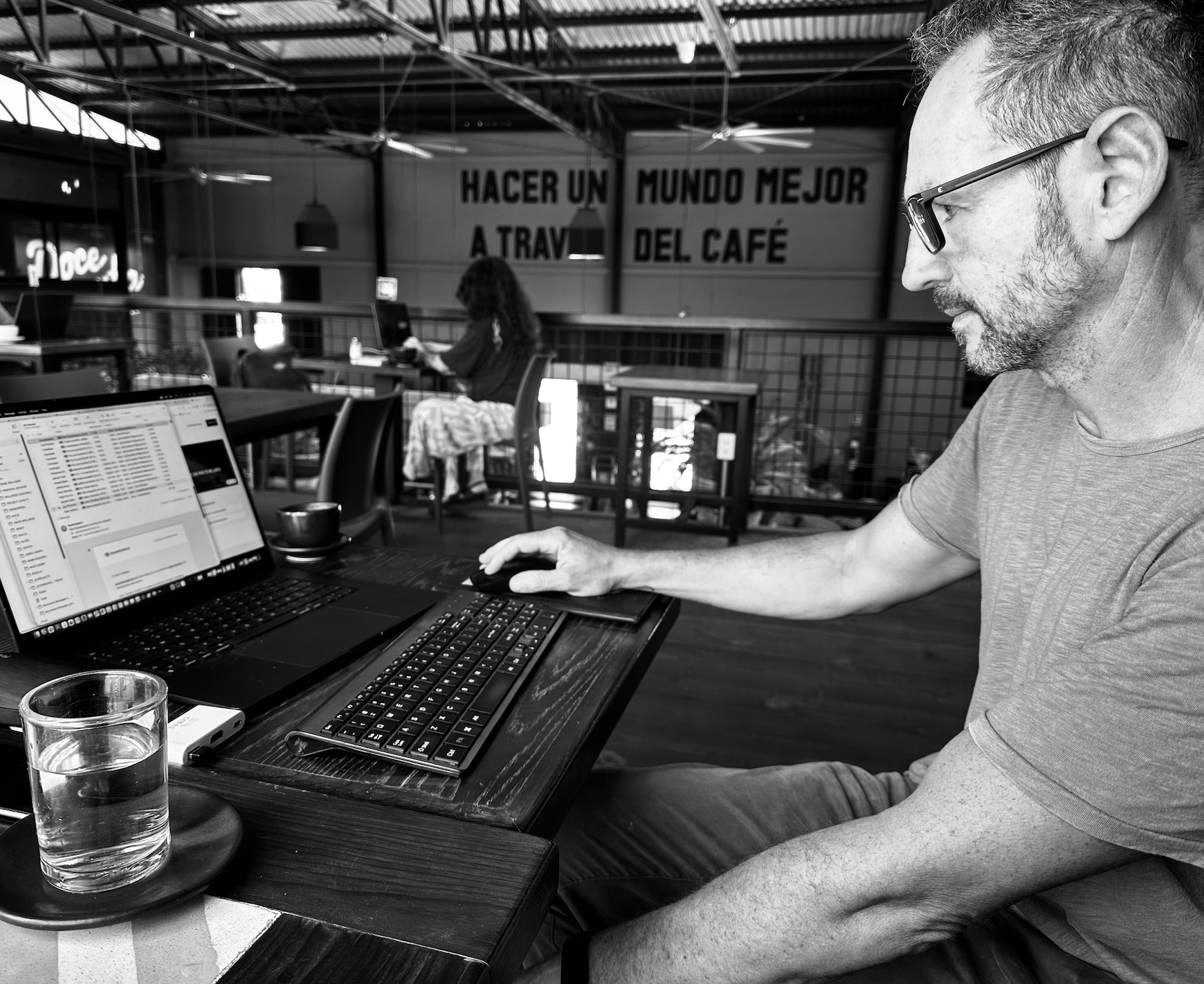 A man with glasses and a beard working on a laptop at a wooden table in a coffee shop. In front of him are a keyboard, mouse, a glass of water on a coaster, and a smartphone. In the background, a woman is sitting at another table working on a laptop, with a sign on the wall that reads 'Hacer un mundo mejor a traves del cafe' in Spanish.