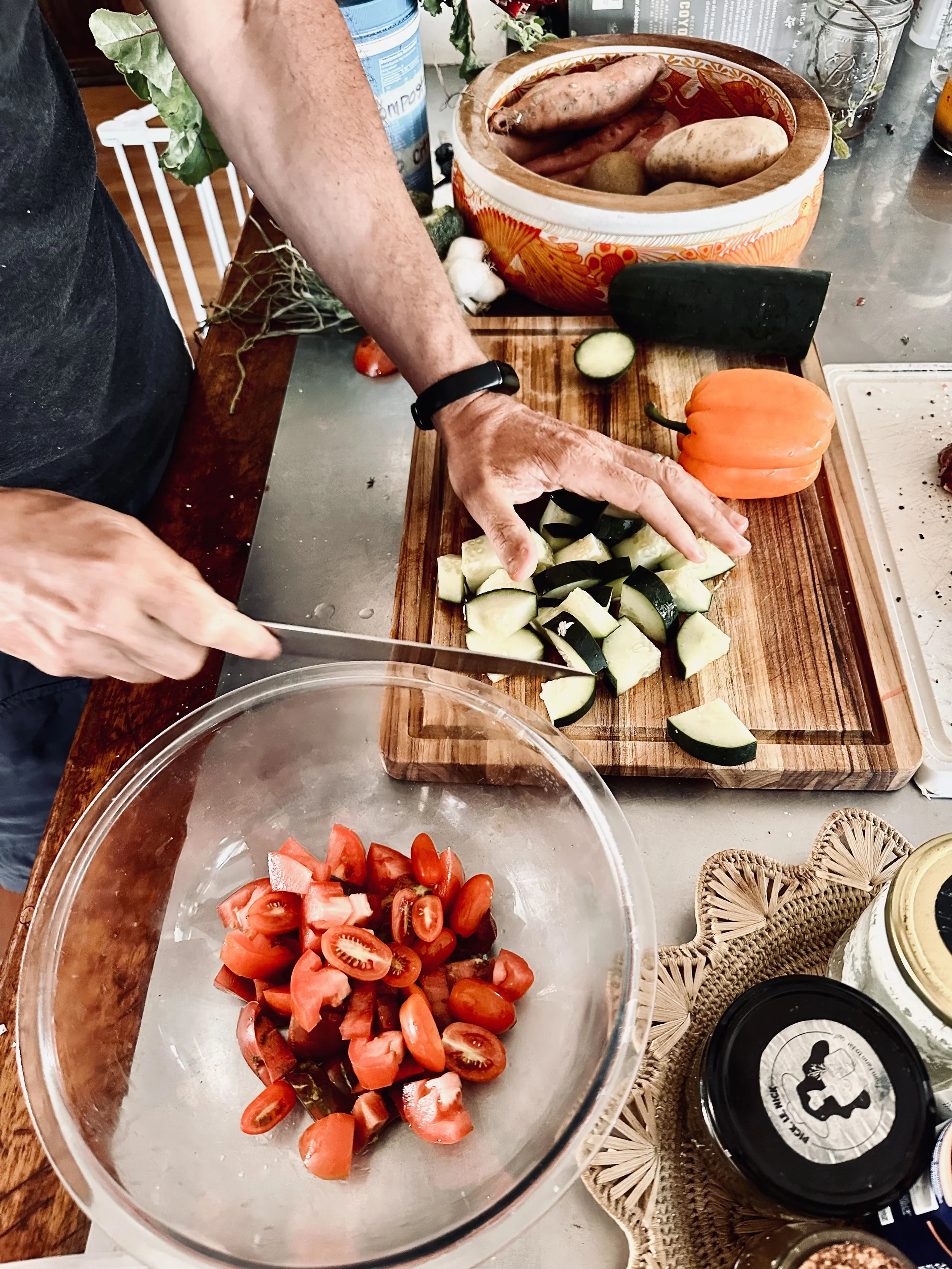 A person chopping cucumbers in a glass bowl, with sliced tomatoes nearby and various vegetables on a wooden cutting board and in a basket. The scene is set on a kitchen counter.