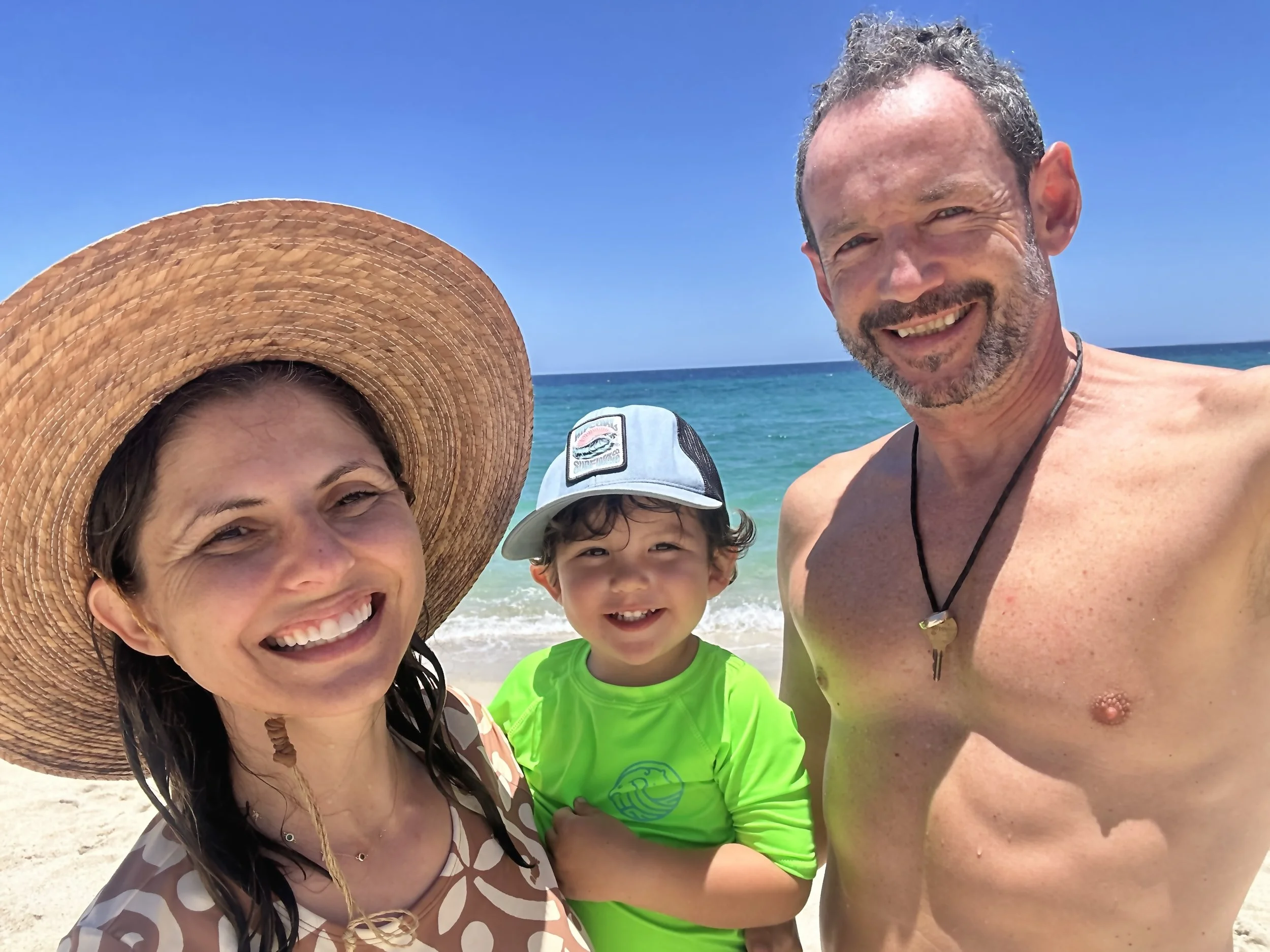 A smiling woman with dark hair wearing a large sun hat, a young boy in bright green shirt, and a smiling man at the beach with the ocean in the background.