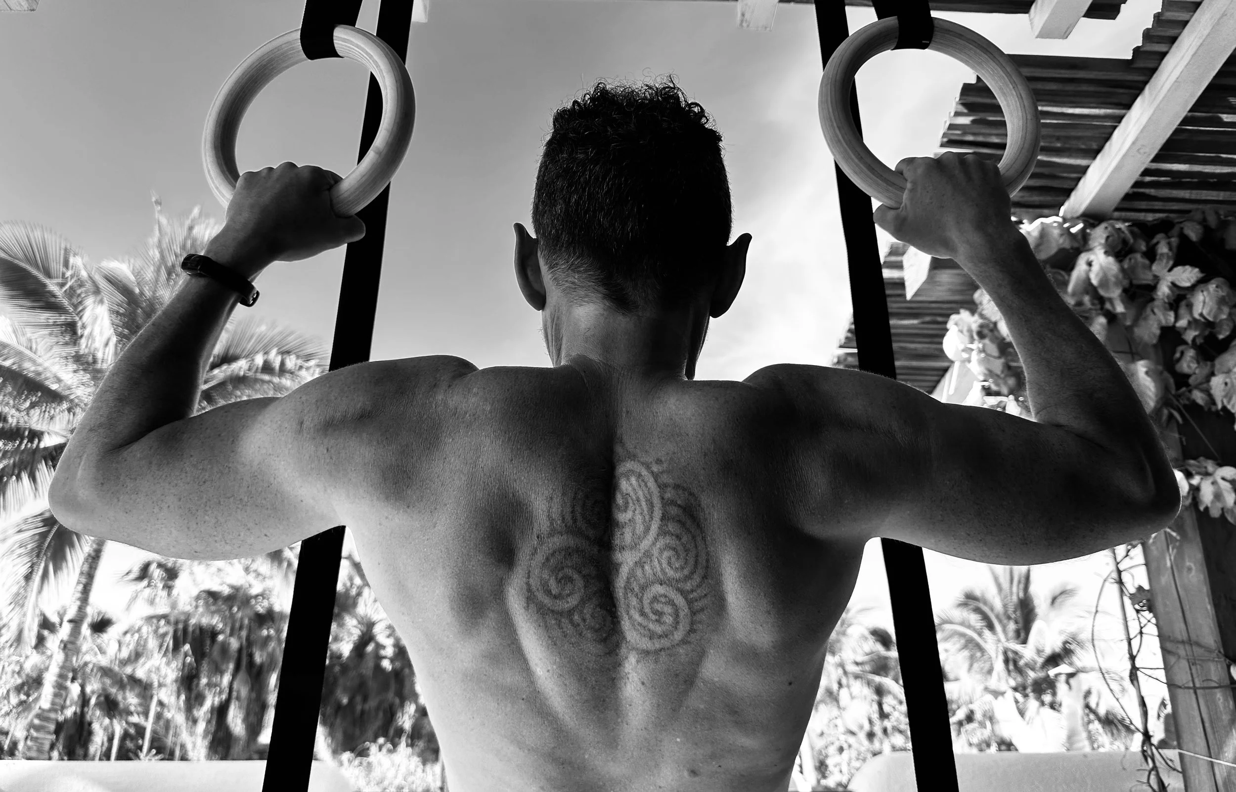 A black and white photo of a man doing pull-ups on gymnastic rings outdoors, viewed from behind, with palm trees and a wooden structure in the background.