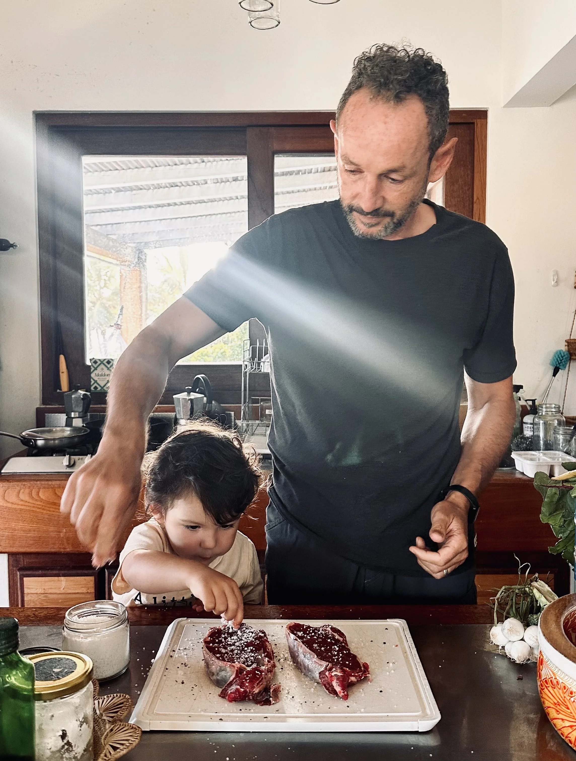 A man and a young girl preparing raw steak for cooking in a kitchen, with the girl reaching for seasoning on a tray, and various kitchen items including garlic, jars, and vegetables visible on the counter.