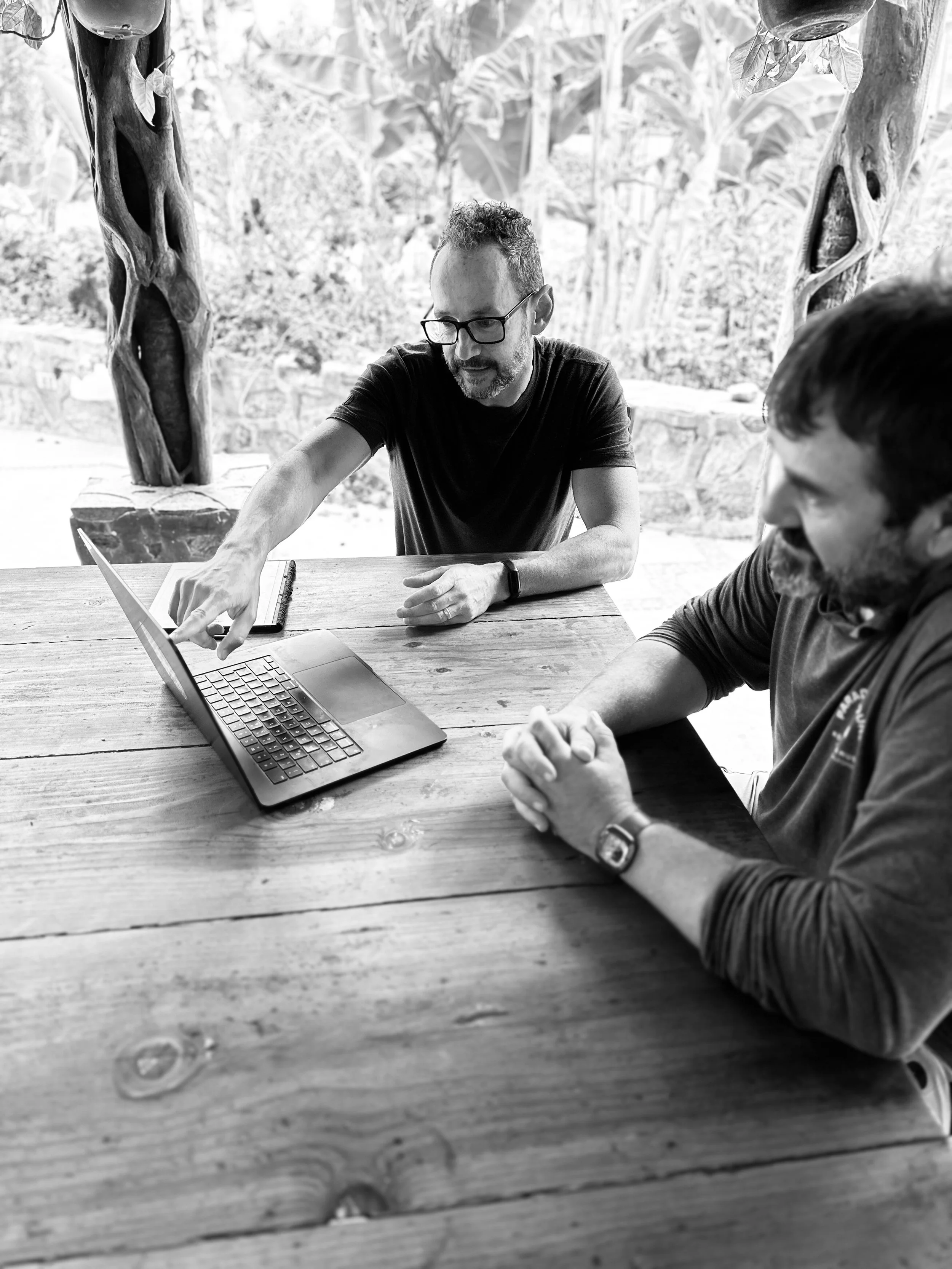 Two men sitting at a wooden table engaged in a discussion, with a laptop open in front of them. One man, wearing glasses and a T-shirt, is pointing at the laptop screen while the other man, also wearing a dark shirt and a wristwatch, looks on. The background features trees and outdoor scenery.