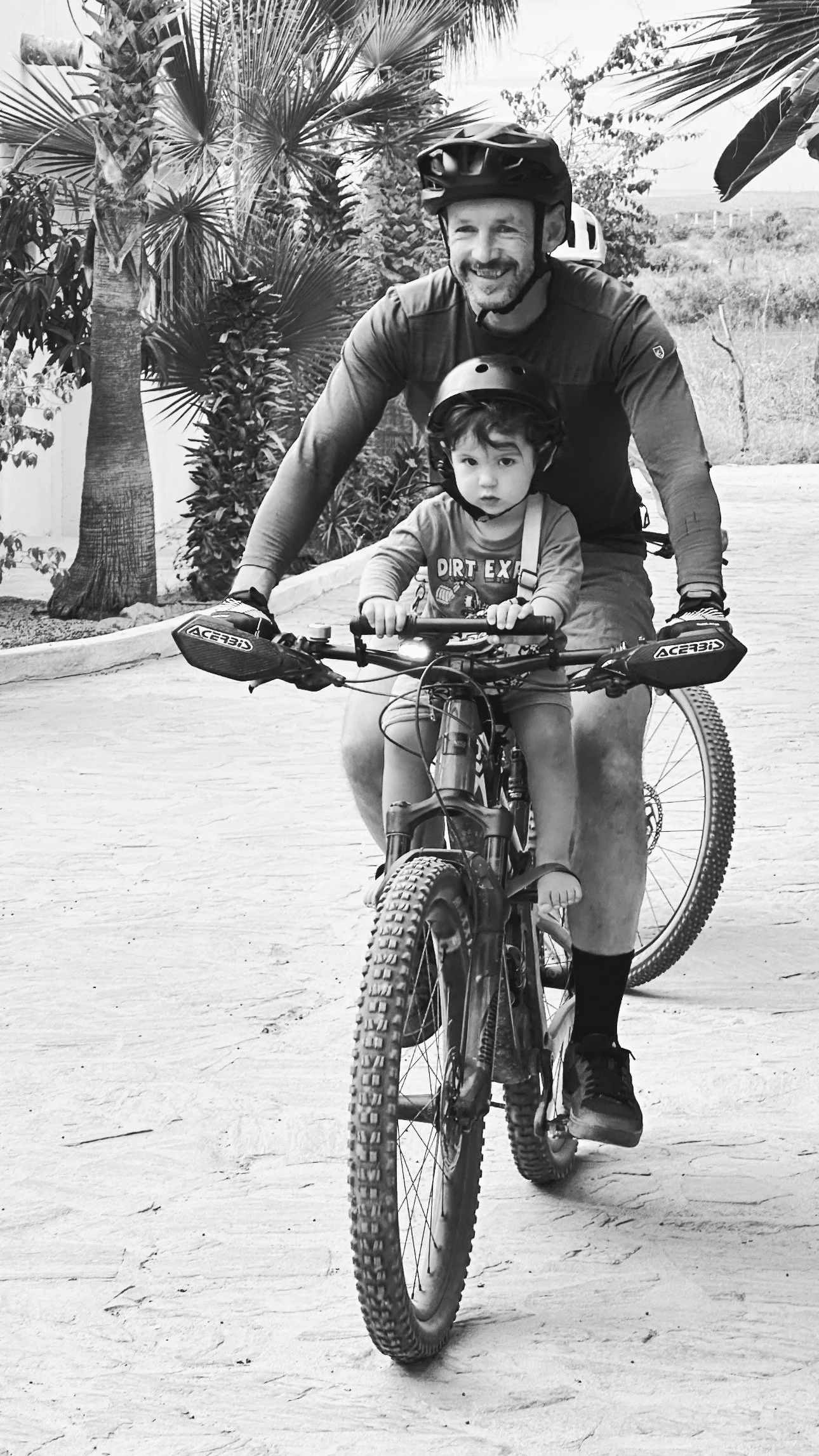 A man and a young boy riding a mountain bike together outdoors, both wearing helmets, with trees and open land in the background. The photo is in black and white.