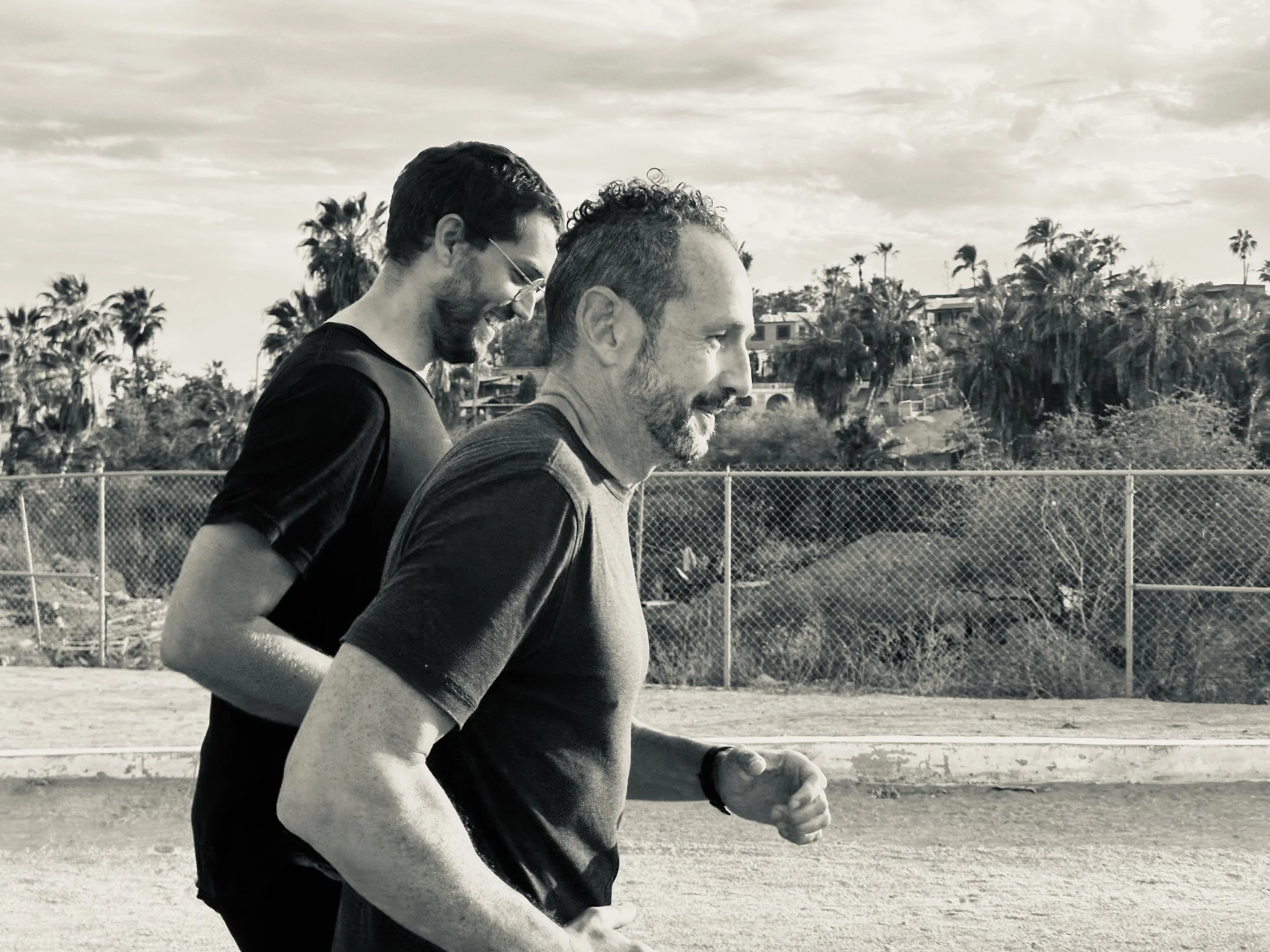 Two men jogging on a track, with palm trees and houses in the background, under partly cloudy sky.