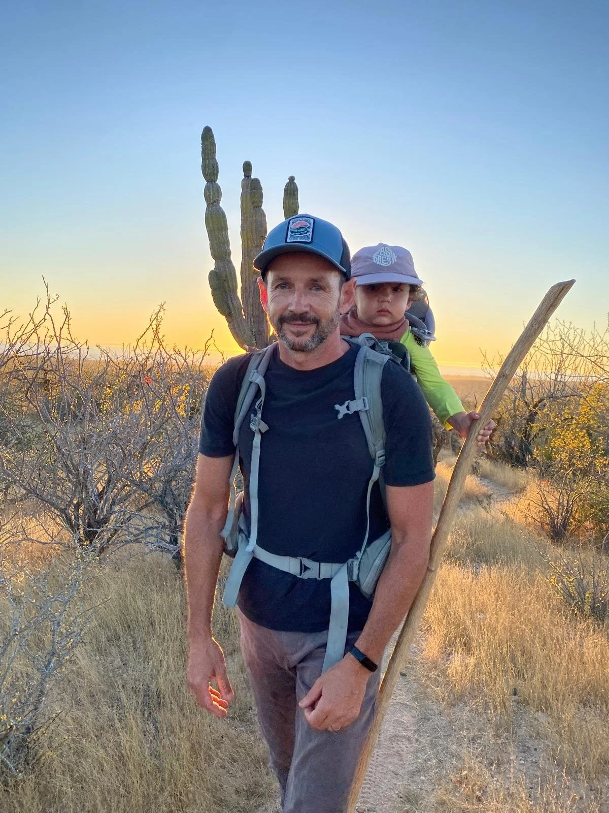 A man with a child on his back hiking in a desert landscape during sunset, with cactus plants and dry bushes around.