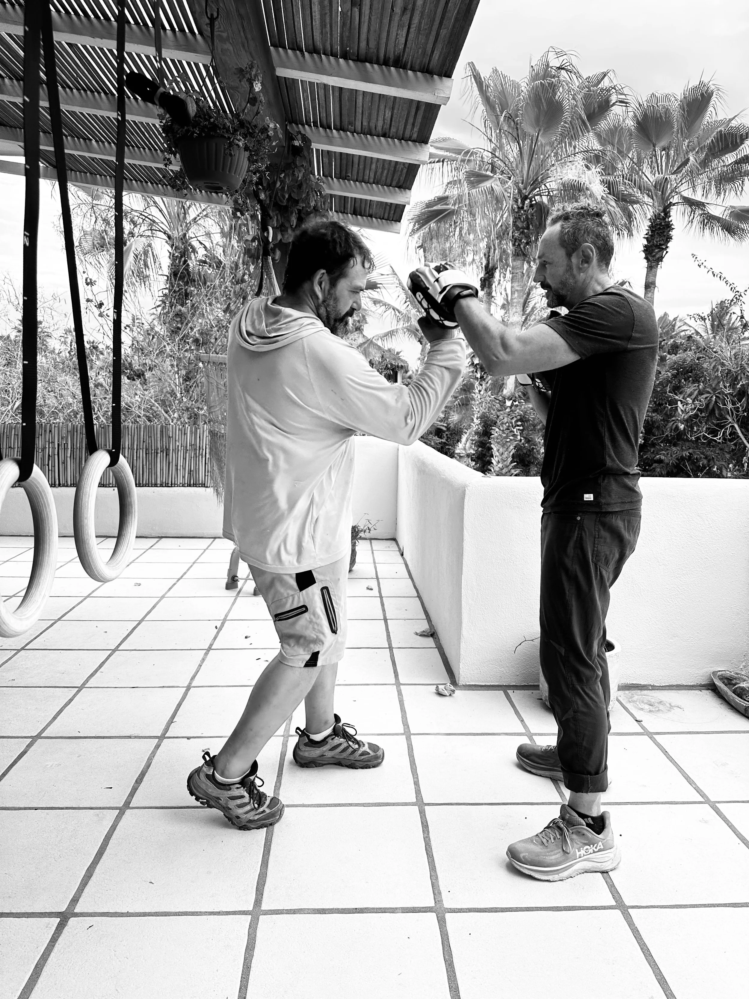 Two men practicing boxing outdoors on a tiled patio, with palm trees and plants in the background. One man is wearing a long sleeve shirt and shorts, the other is wearing a t-shirt and pants. They are facing each other with gloves on, engaged in a boxing training session.