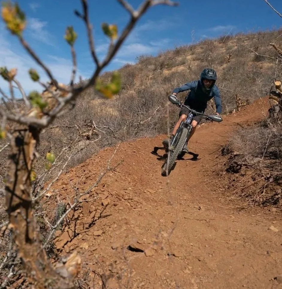 A mountain biker riding down a dirt trail on a dry hillside with sparse bushes and a blue sky.