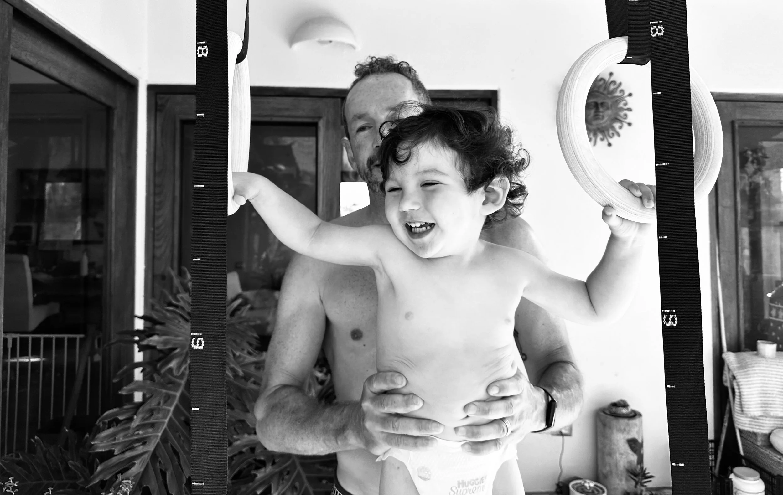 A young boy with curly hair smiling and holding on to gymnastic rings, with a man behind him lifting him up. The background shows a room with wooden furniture and a decorative wall hanging.
