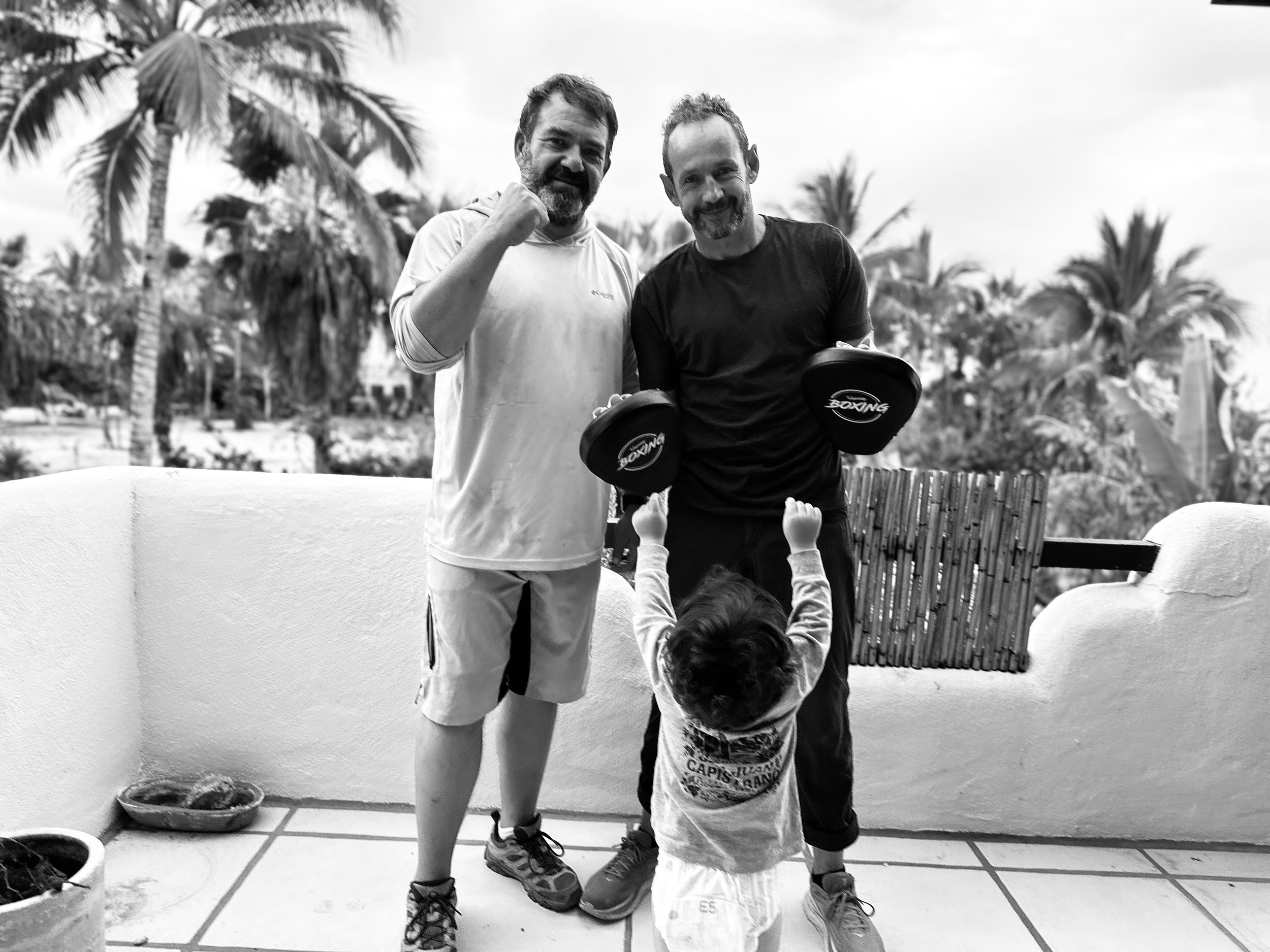 Two adult men and two children outdoors surrounded by palm trees. The men are smiling; one has a beard and is wearing a light shirt, the other is clean-shaven and wearing a dark shirt. One child is holding boxing gloves, and the other is partly visible at the bottom of the image.