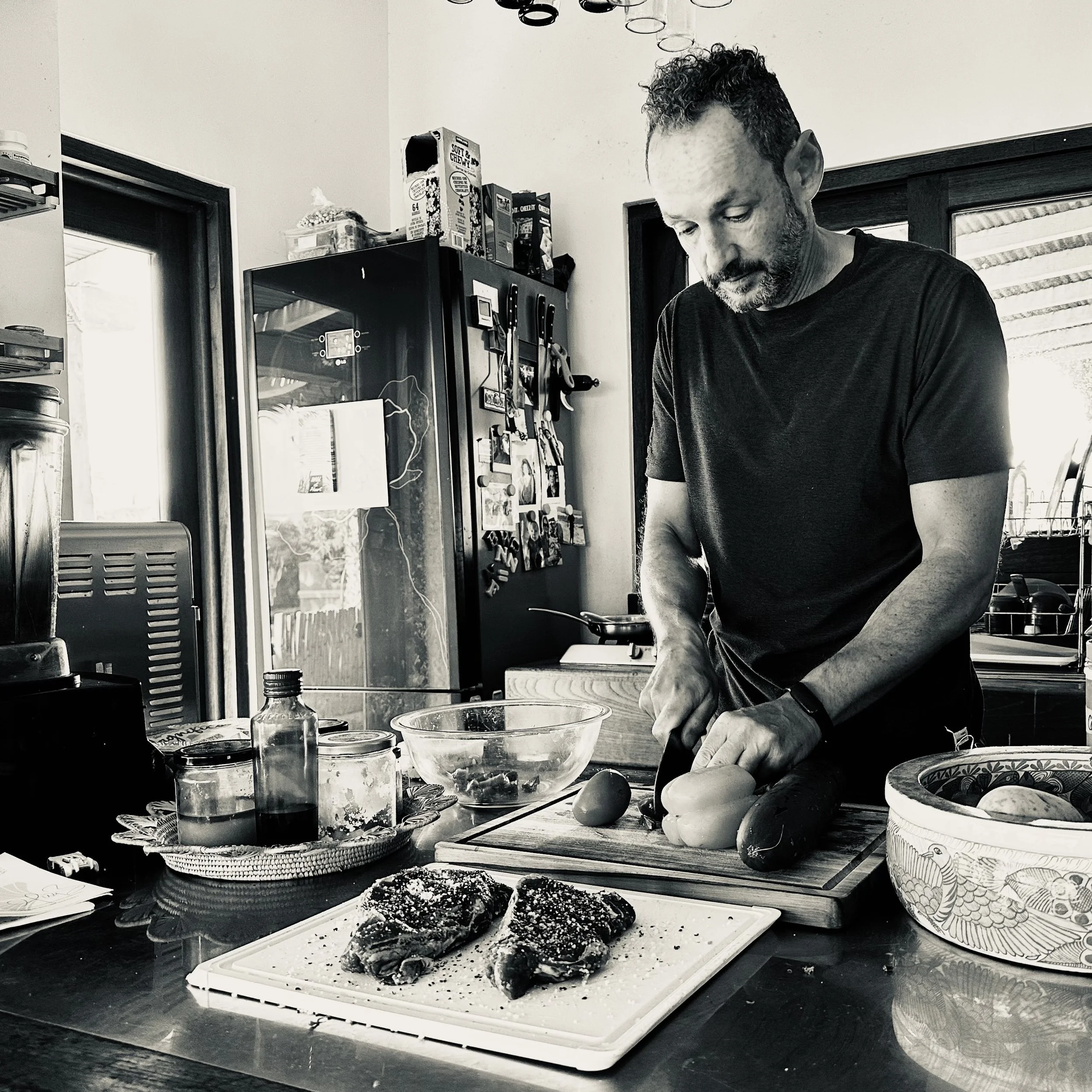 A man with curly hair and a beard preparing food in a kitchen. He is slicing a vegetable on a cutting board. The kitchen has a variety of containers, jars, and appliances on the counter. There are decorative plates and utensils on the wall. The image is in black and white.