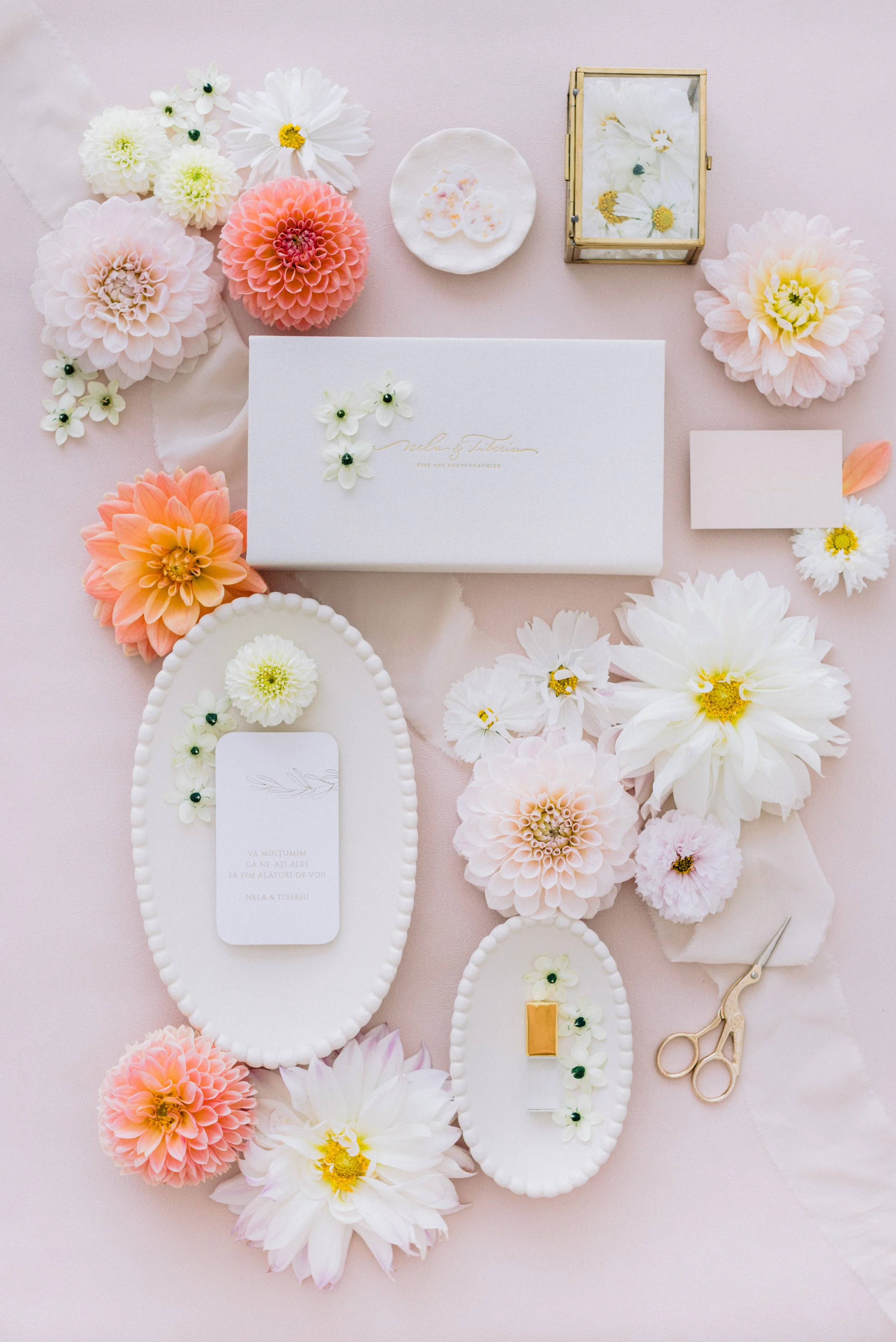 Aranjament de flori, inclusiv dalii, margarete si alte flori, pe un fundal roz deschis cu invitatii de nunta, mici obiecte decorative, o foarfeca aurie si diverse tablouri inramate cu flori, intr-un flat lay realizat de Nela.