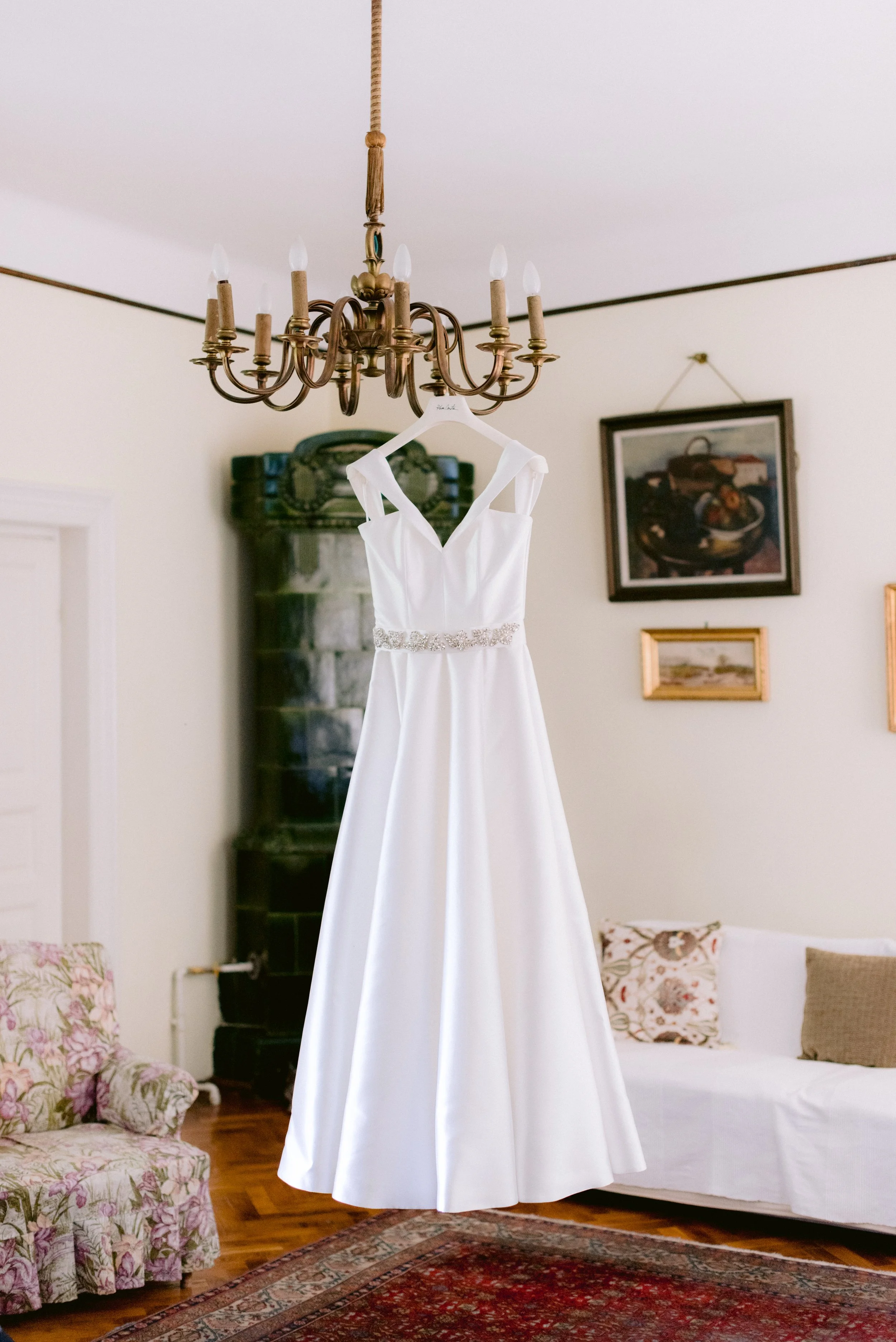 Rochie de mireasa alba agatata de un candelabru intr-un living cu mobilier vintage si tablouri inramate.