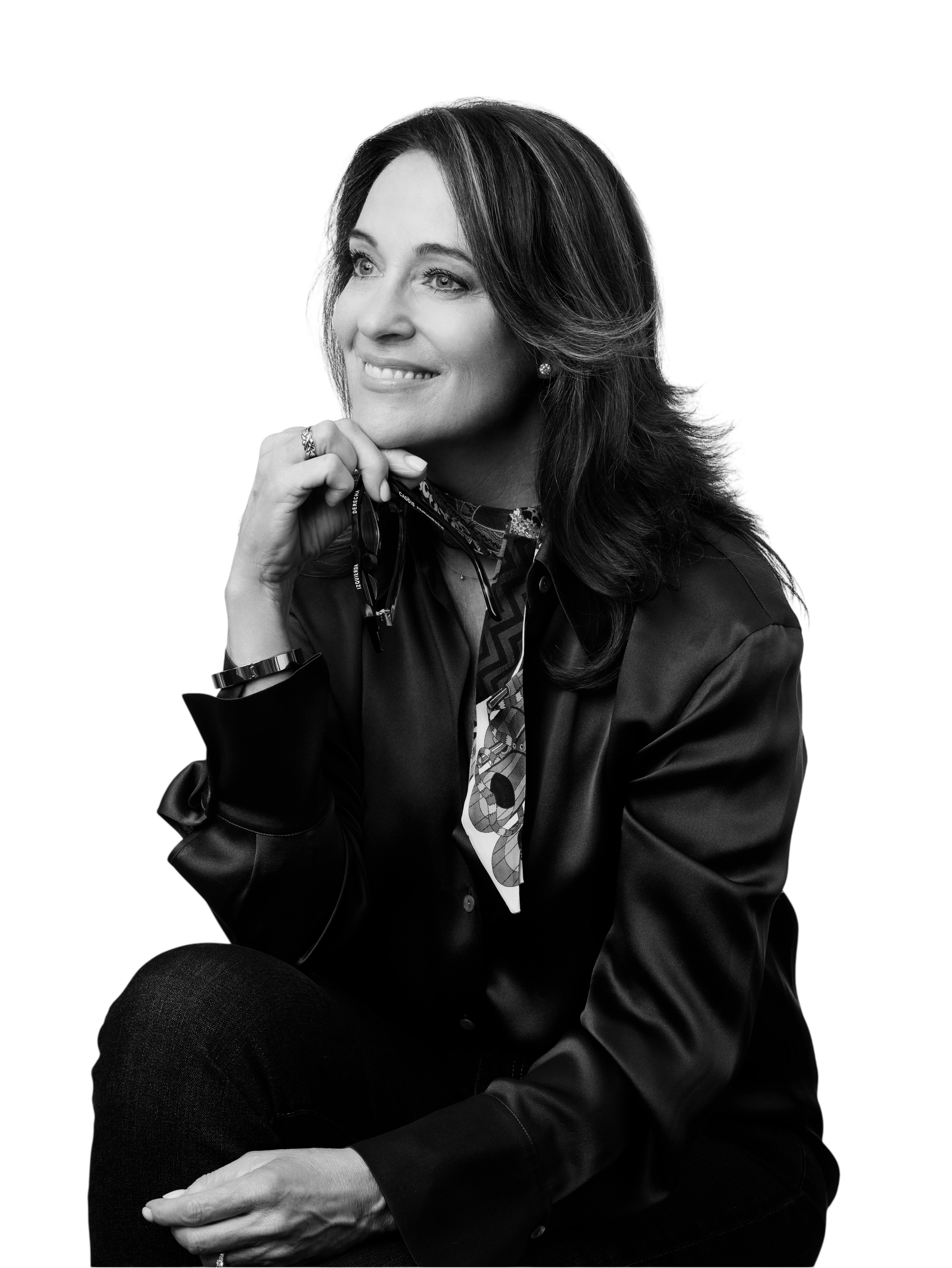 Black and white photo of mktgcollab founder with shoulder-length hair, smiling and looking to her left, sitting with her hand resting on her knee, wearing a dark jacket, patterned scarf, watch, and earrings.