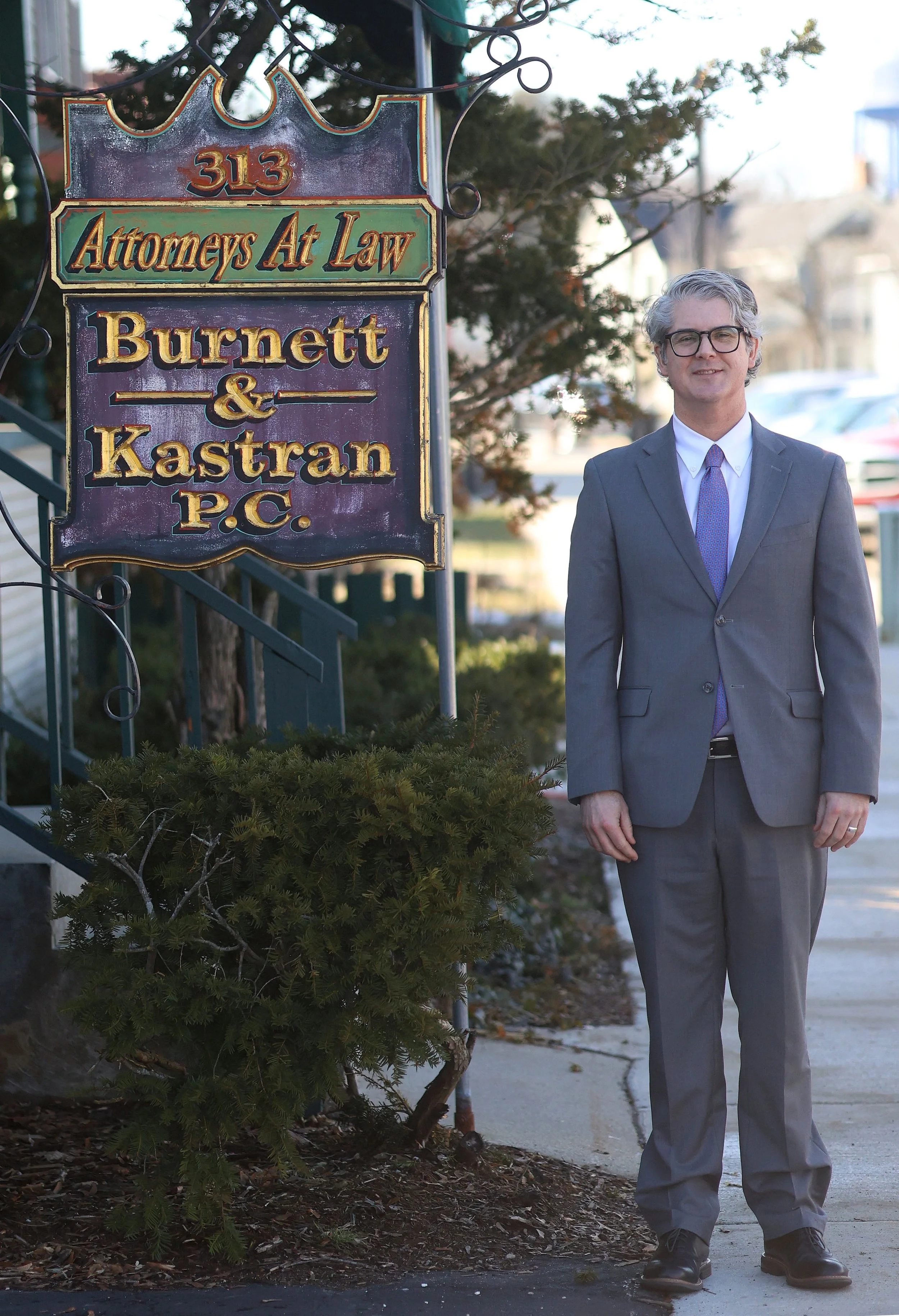 A man in a gray suit and glasses standing outside next to a sign that reads 'Attorneys At Law Burnett & Kastran P.C.' with a tree and buildings in the background.
