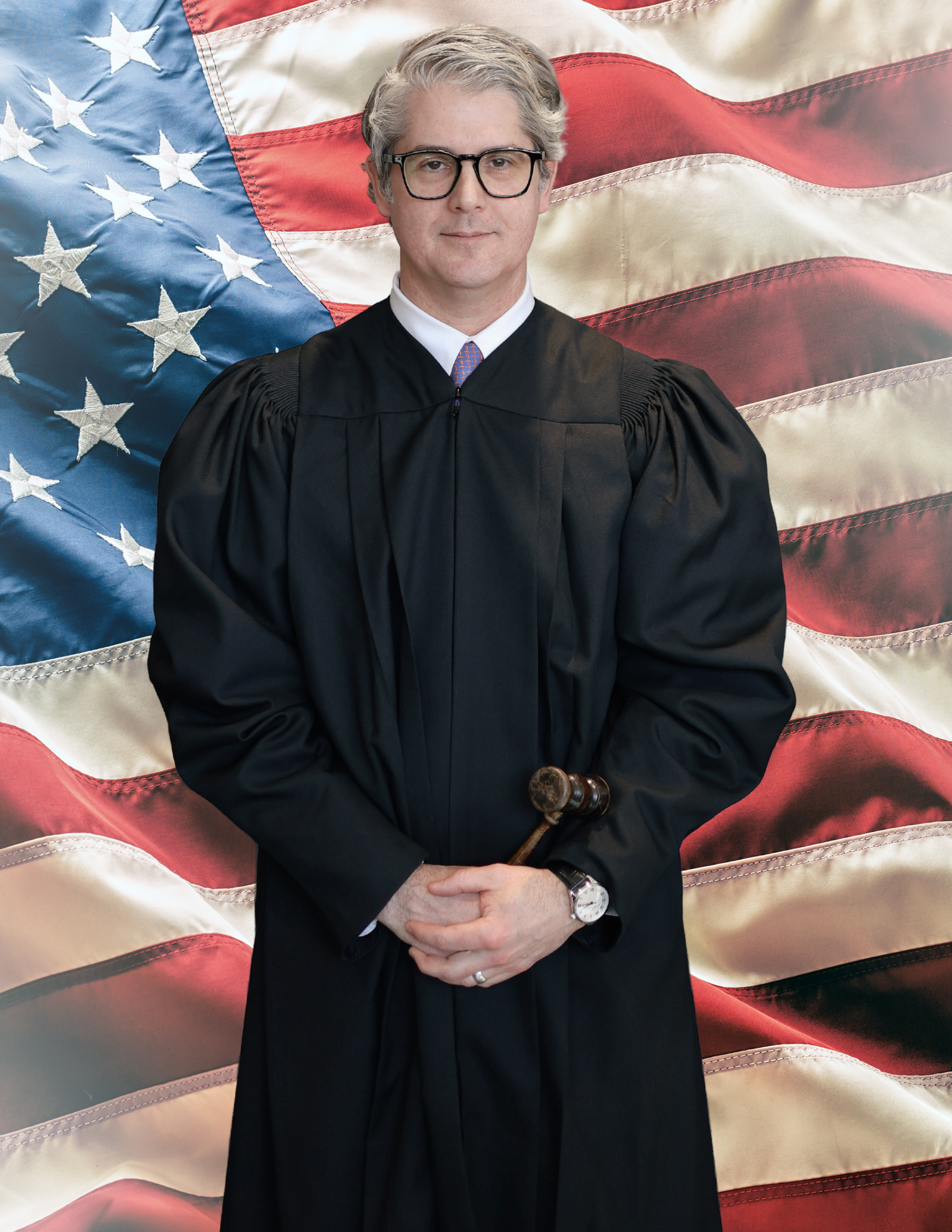 A judge in black robes holding a gavel, standing in front of an American flag.