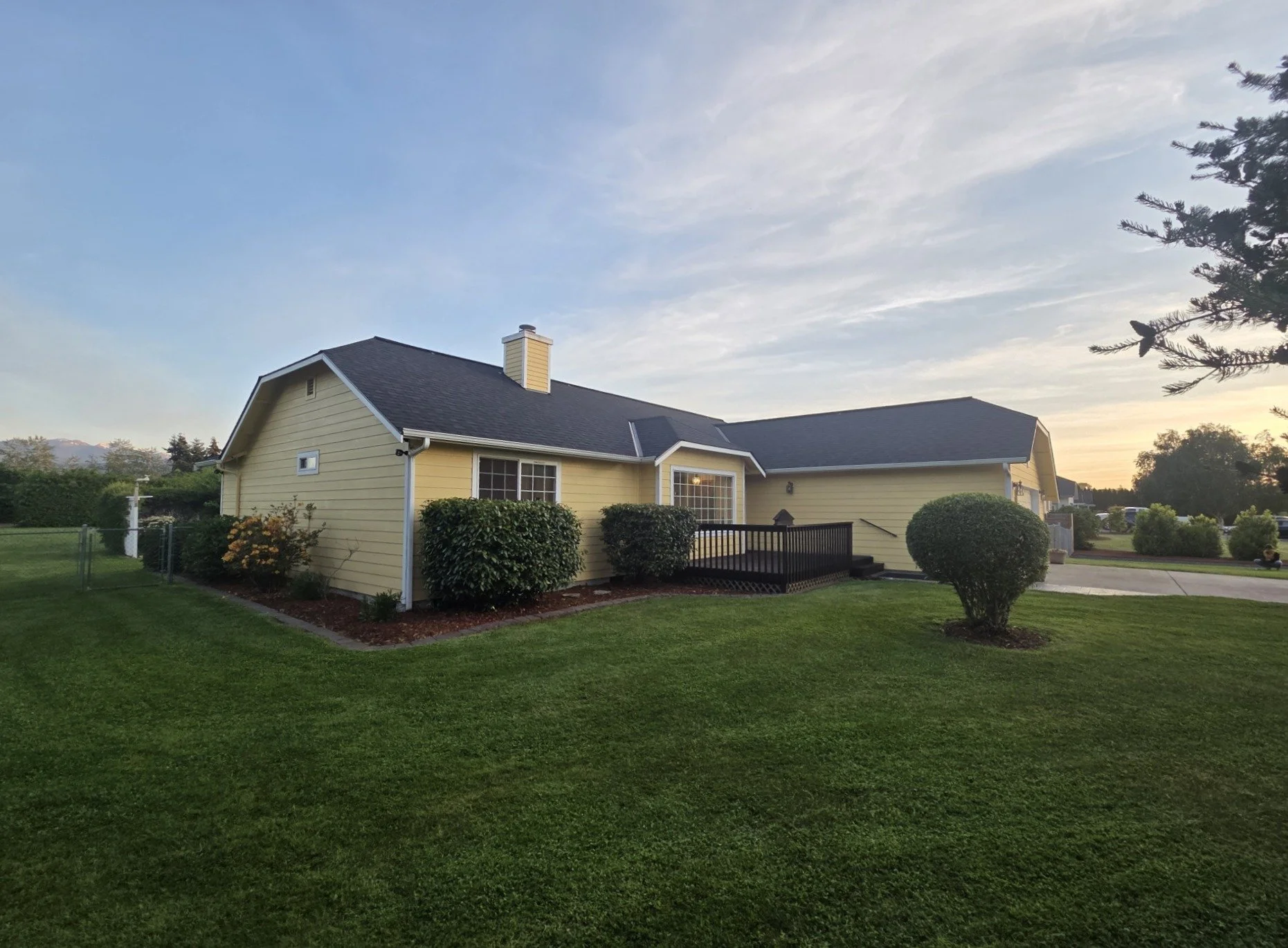 A yellow single-story house with a dark roof, surrounded by a well-maintained lawn, bushes, trees, and a paving walkway, with a sky showing some clouds at sunset.