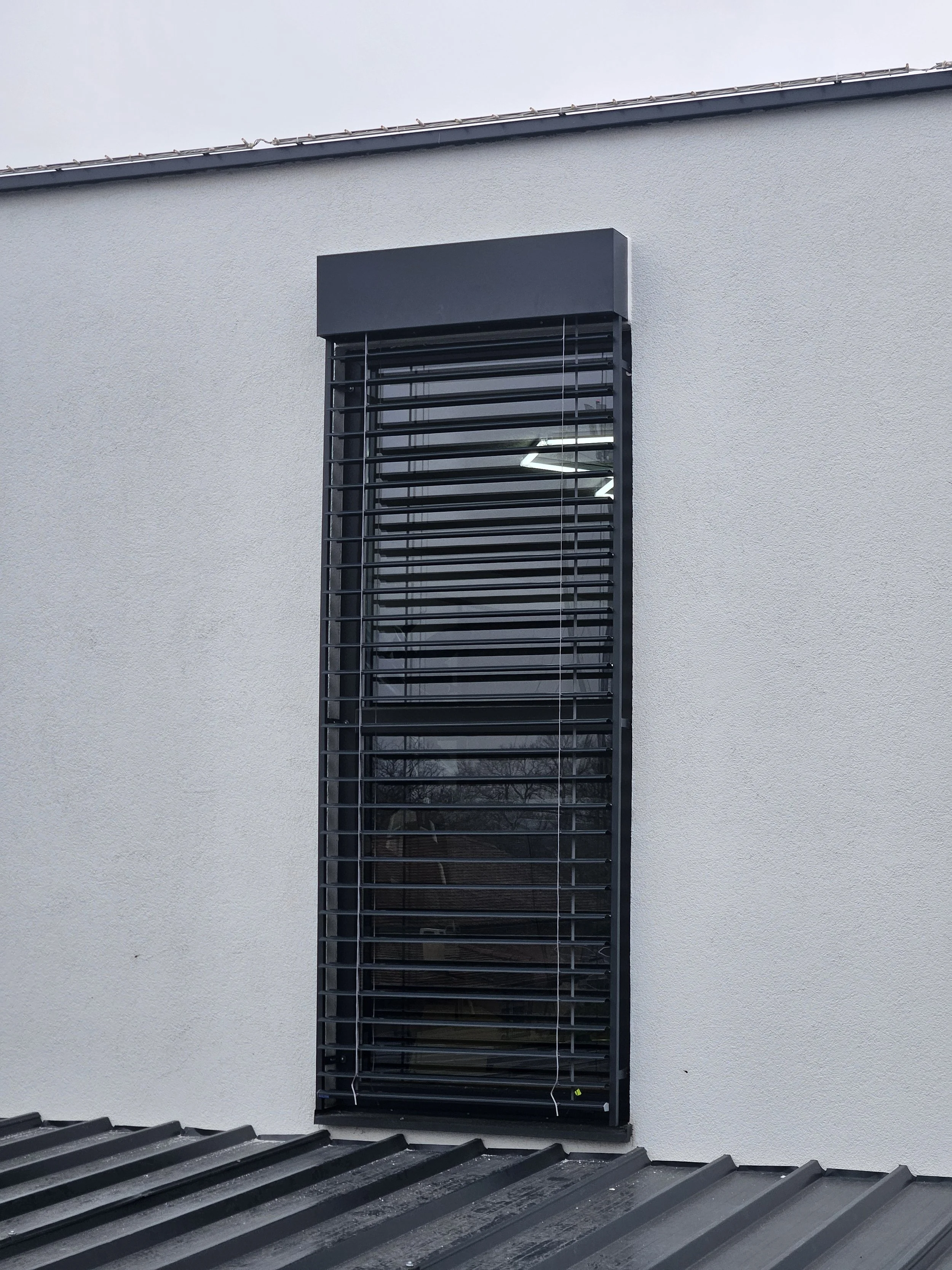 Exterior window with dark horizontal blinds on a white stucco building above a corrugated metal roof