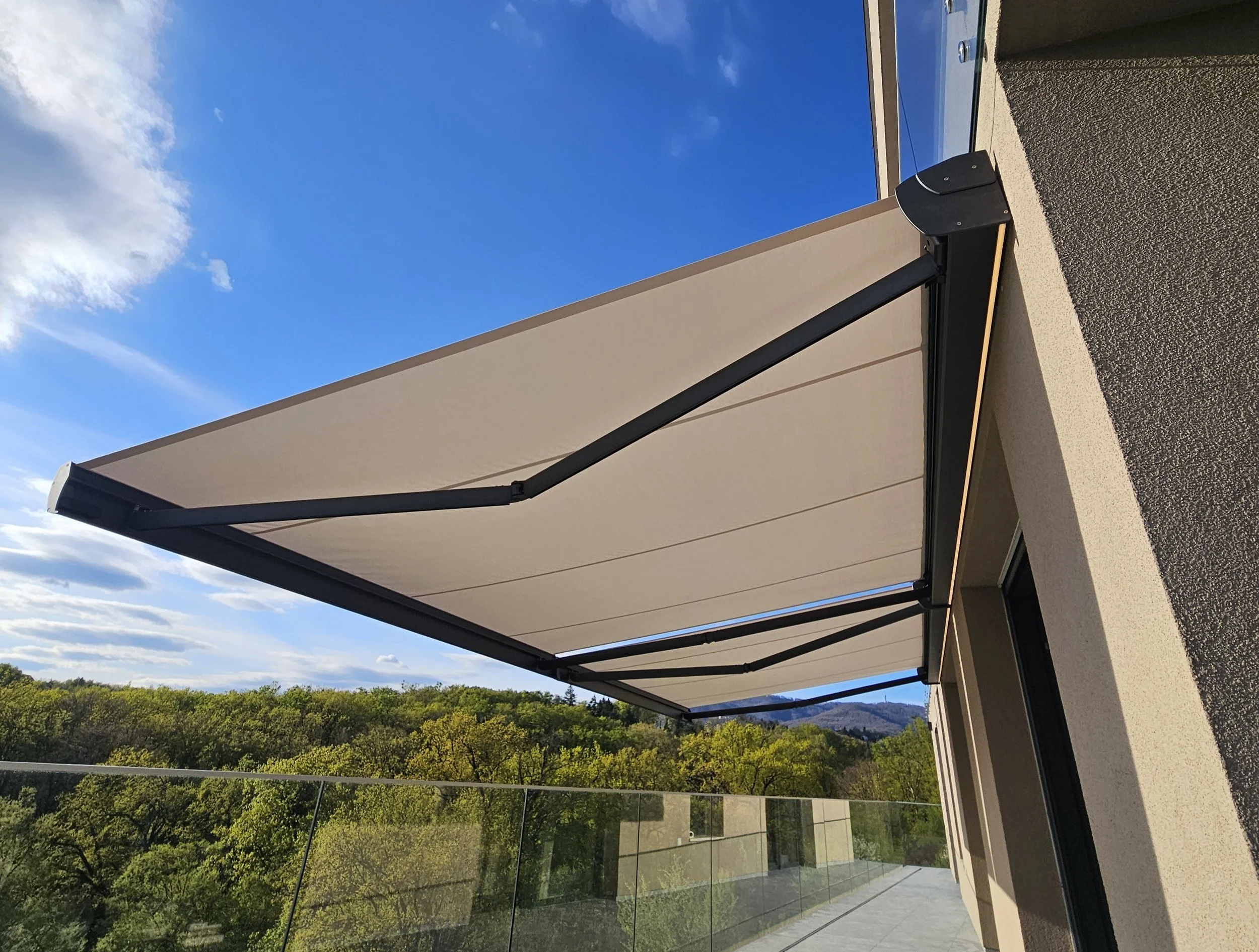 Retractable awning shading a modern balcony with glass railing, green trees, and distant mountains under a blue sky