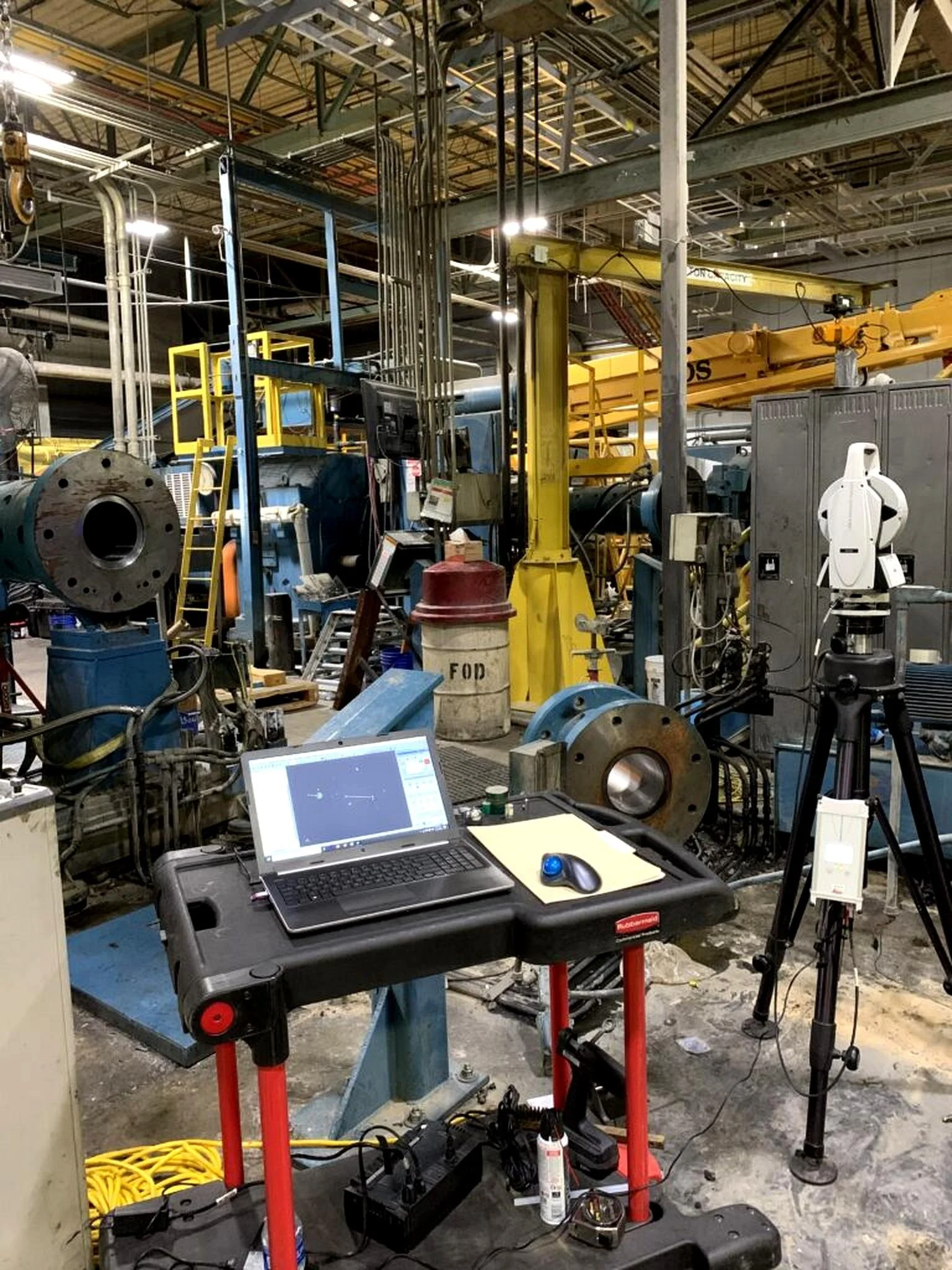 Industrial workshop with machinery, a laptop on a workbench, cables, and tools, with a metal ceiling and exposed pipes.