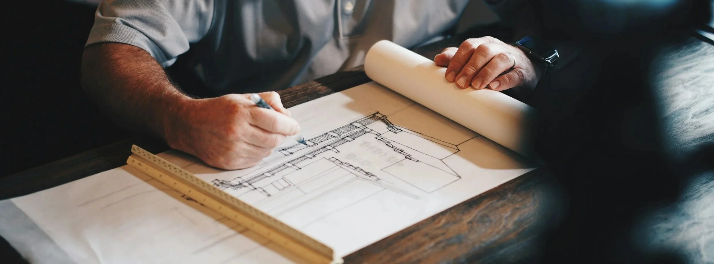 A person drawing an architectural blueprint at a table with a ruler nearby.