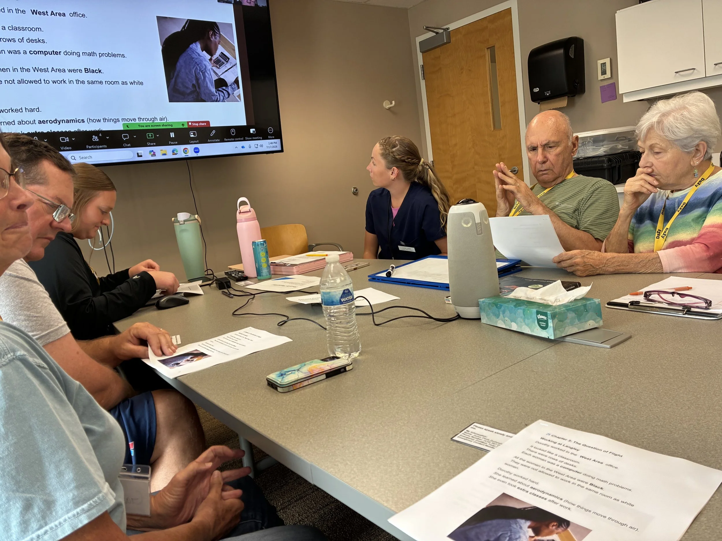 An aphasia group sits around a conference table. 6 people are visible, including people with aphasia, caregivers, and student clinicians. A sliver of a presentation visible in the corner