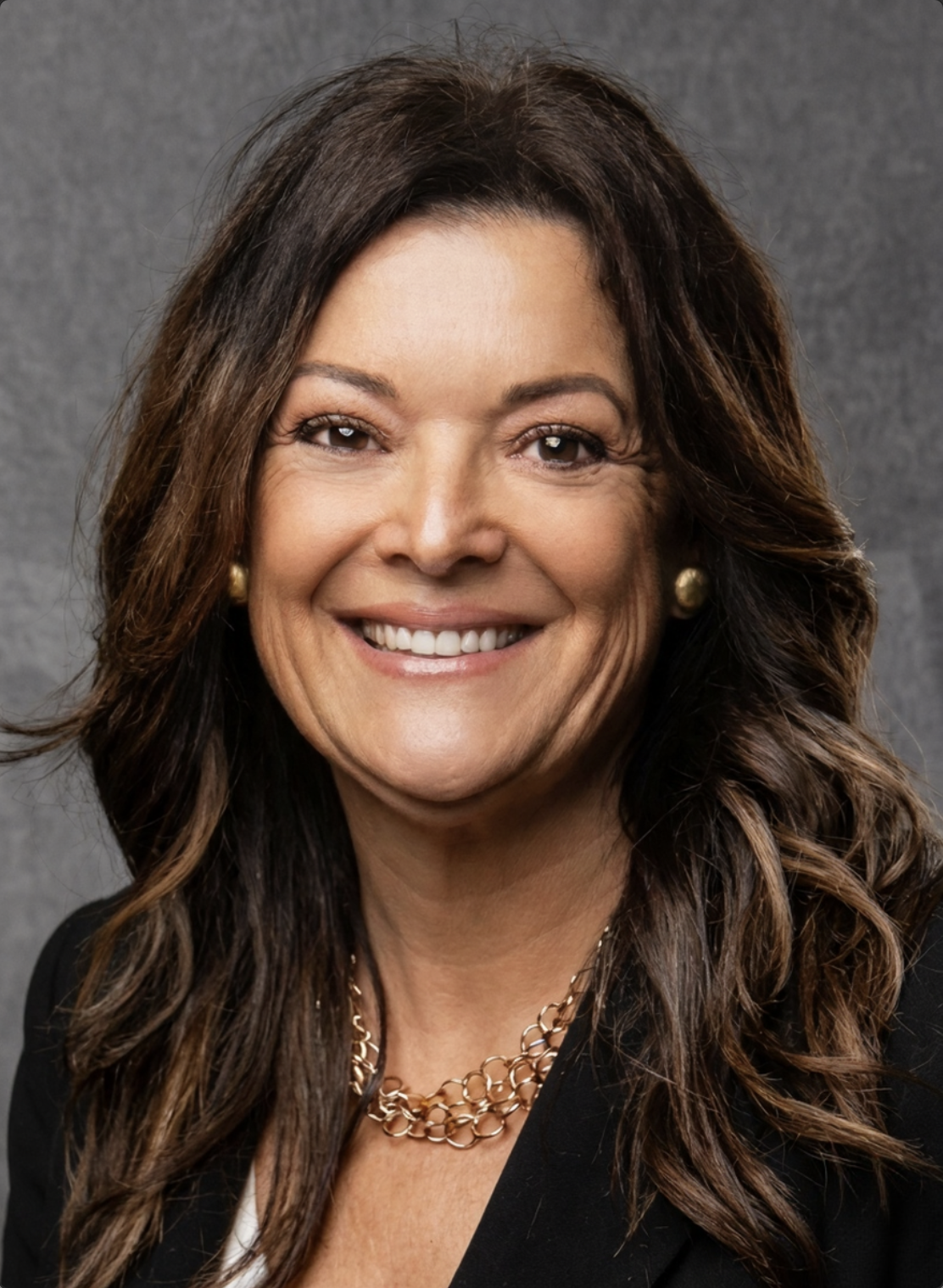 A woman with long, dark brown wavy hair wearing a black blazer, gold earrings, a gold necklace, and smiling against a gray background.