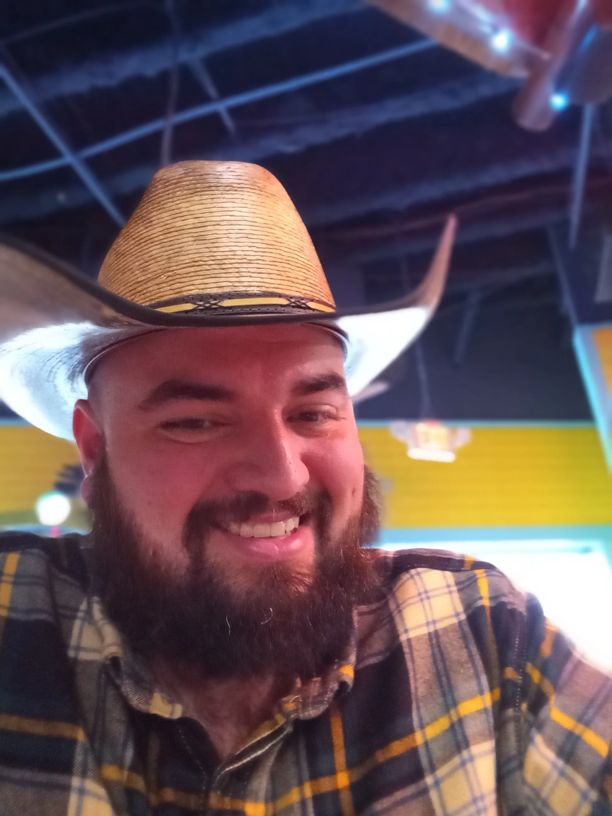 A man with a beard and a big smile wearing a plaid shirt and a large cowboy hat in a festive environment.