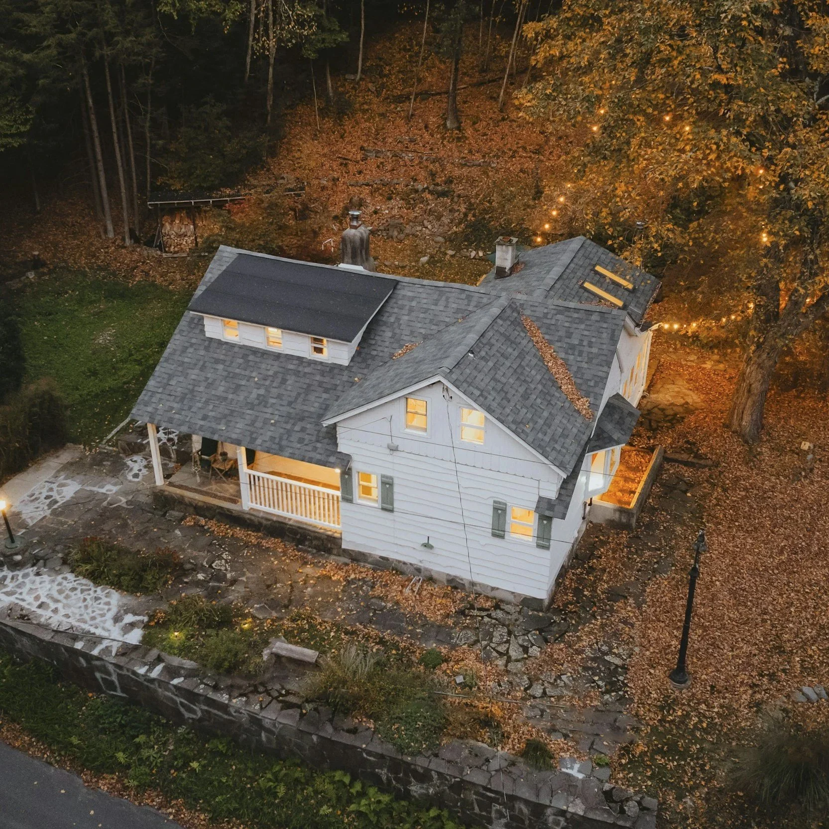 A two-story white house with a gray roof, illuminated by warm outdoor lights, surrounded by autumn leaves and trees at dusk.