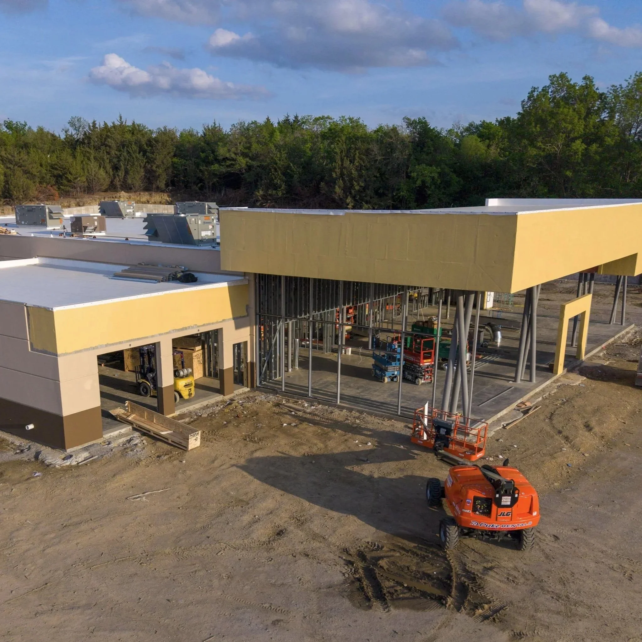 Construction site with a building framework, construction equipment, and a lift, set against a backdrop of trees and a partly cloudy sky.