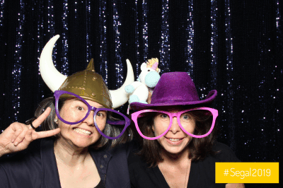 Jessamyn and Jane strike several silly poses wearing giant colourful glasses, party hats, and holding a small crochet unicon