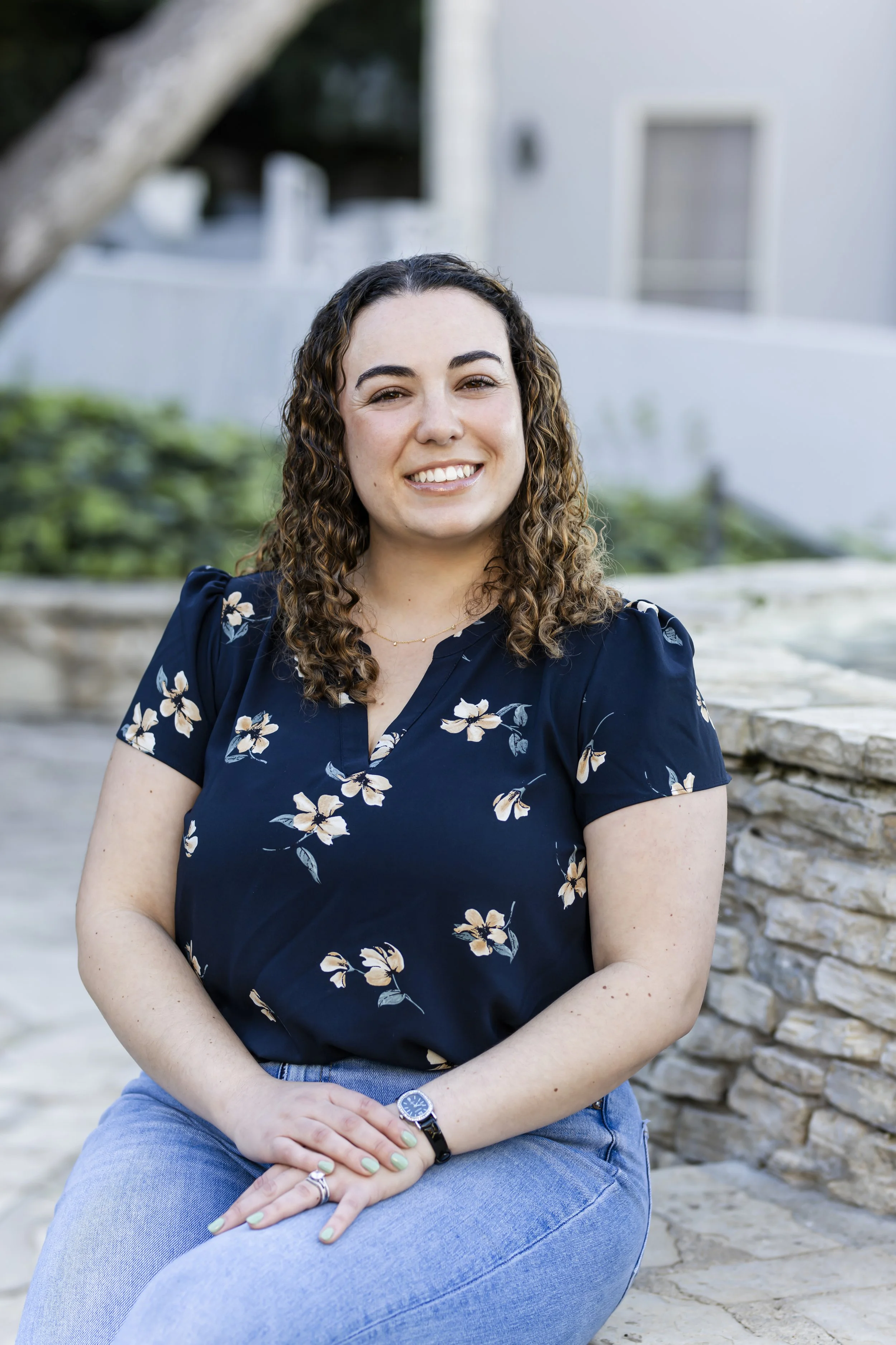 A smiling woman with curly brown hair sitting outdoors on a stone ledge, wearing a navy blue floral top and light blue jeans.