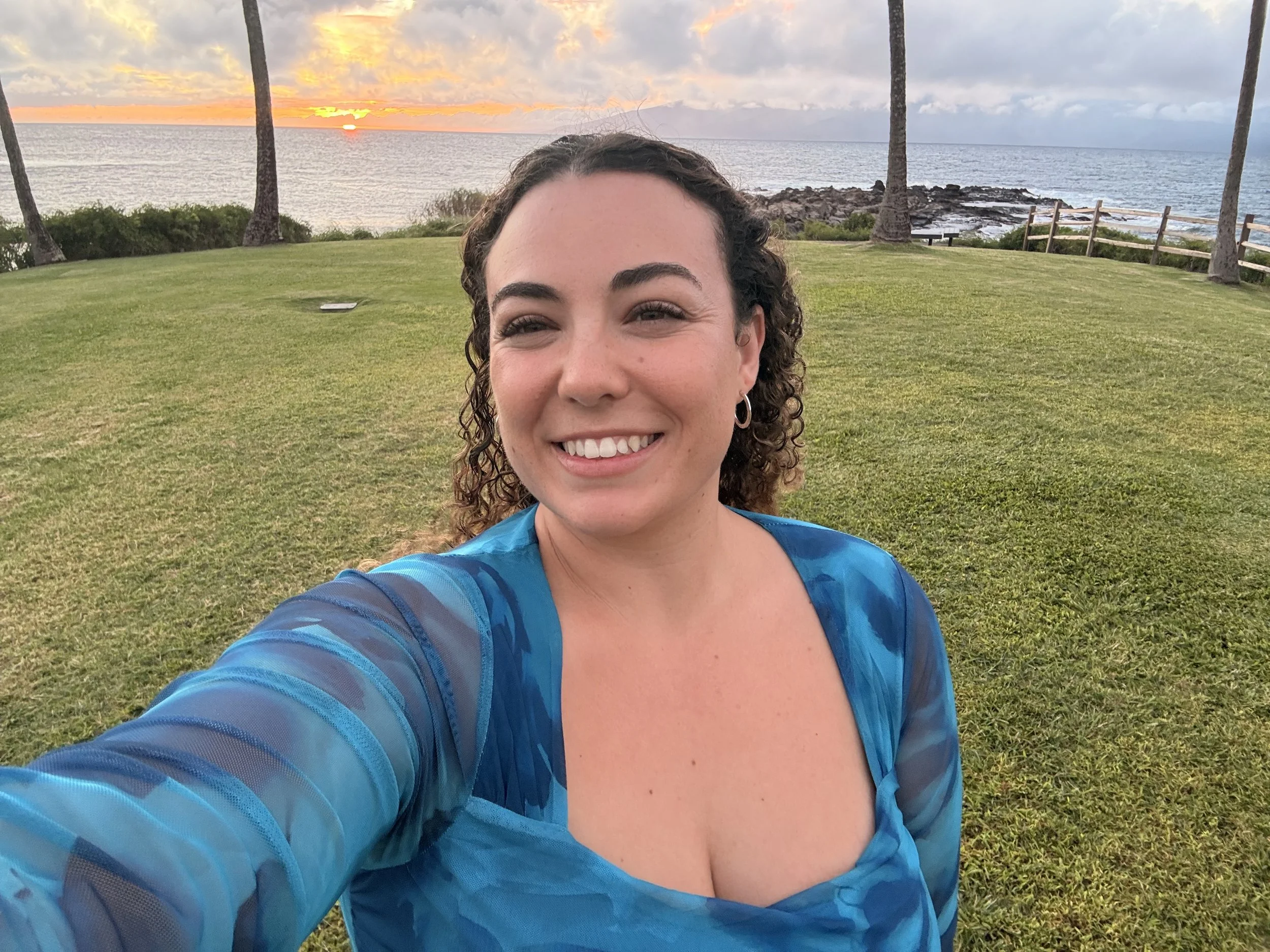 Smiling woman with curly hair taking a selfie on a grassy area overlooking the ocean at sunset, with palm trees and rocks in the background.