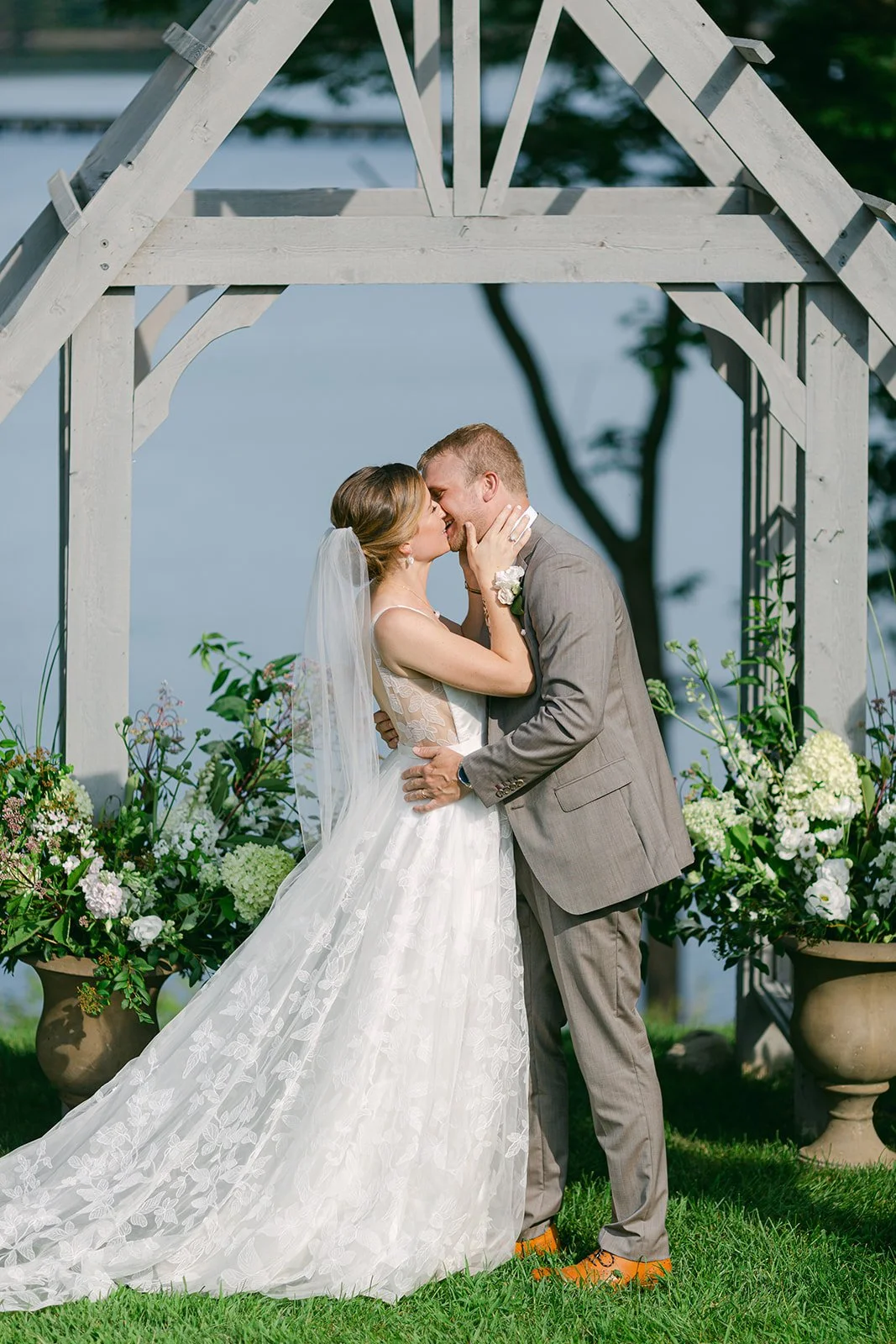 A bride and groom kissing under a wedding arch decorated with flowers, outdoors by a body of water.