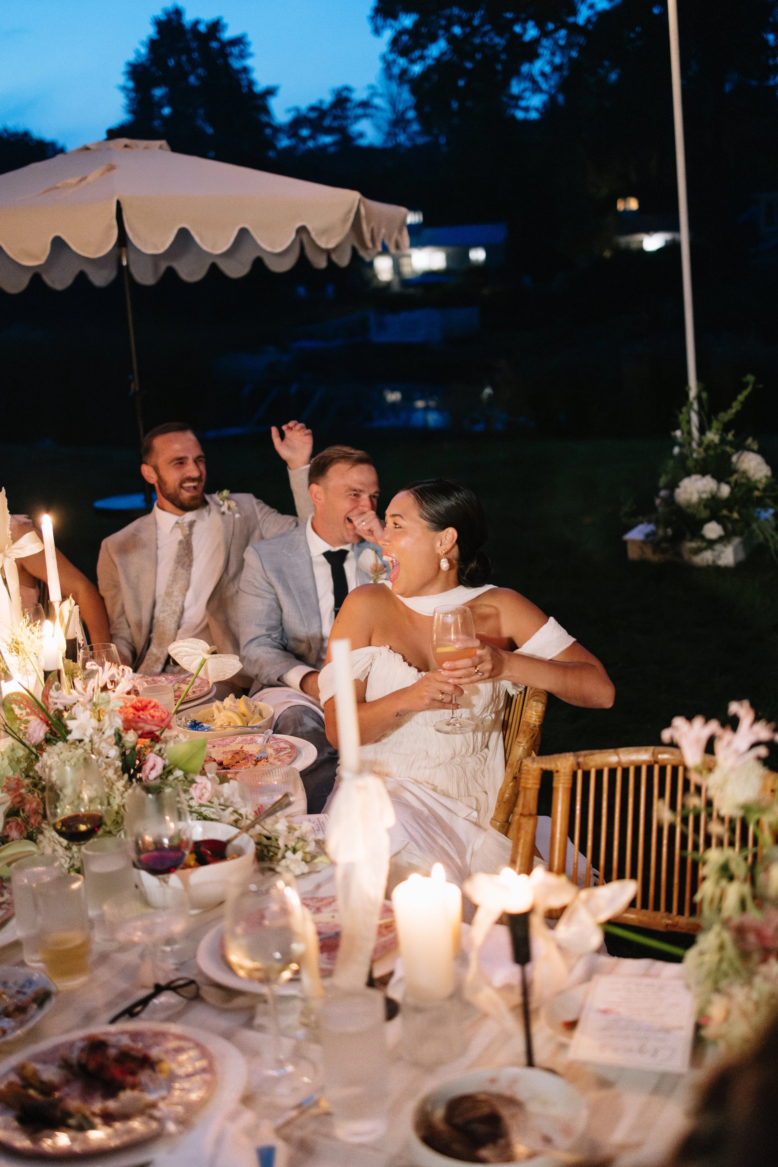 A group of wedding guests celebrating outdoors during the evening, with a decorated table, candles, and floral arrangements, enjoying a joyful moment.