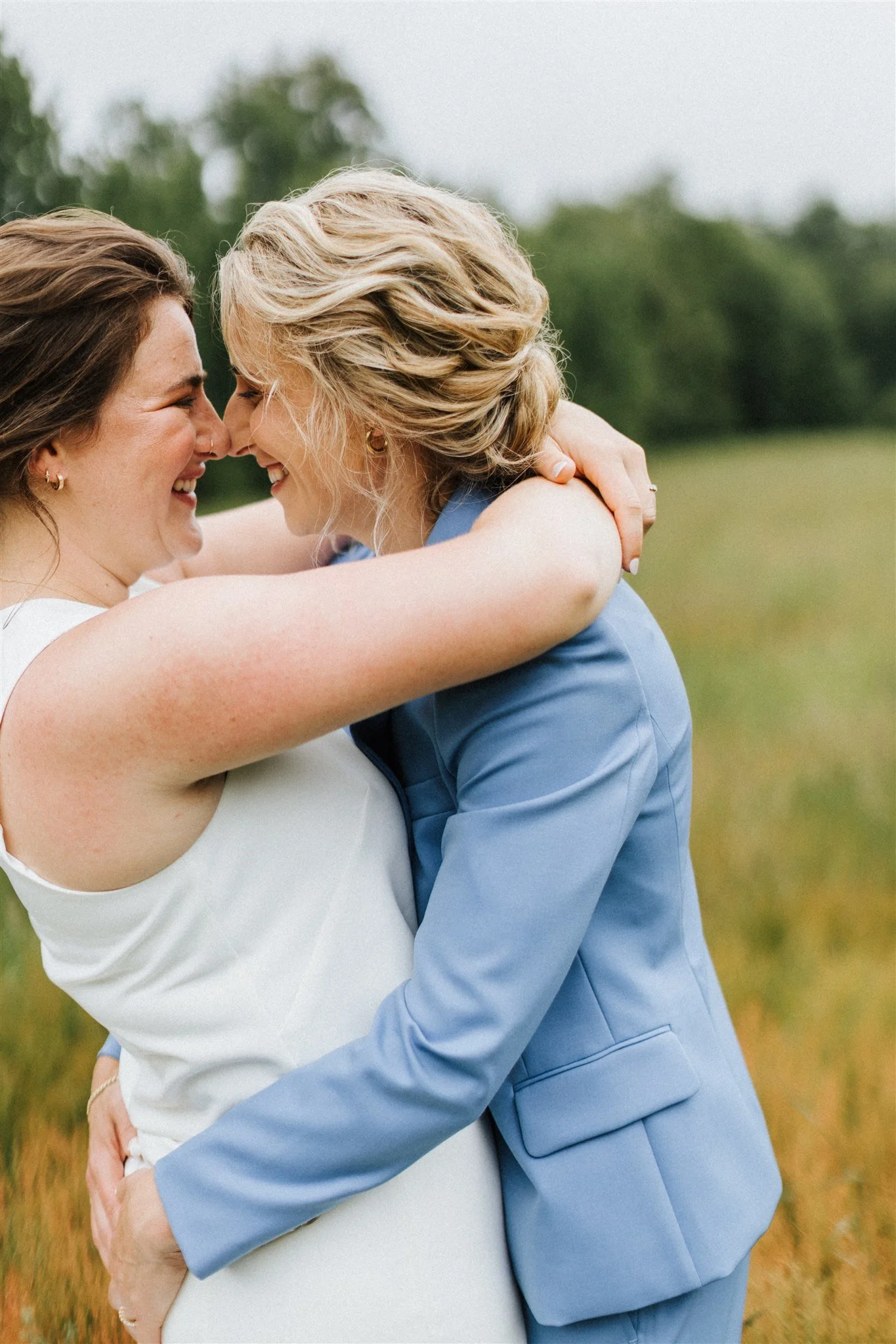 Two women with blonde hair, smiling and embracing each other outdoors in a field with greenery in the background.