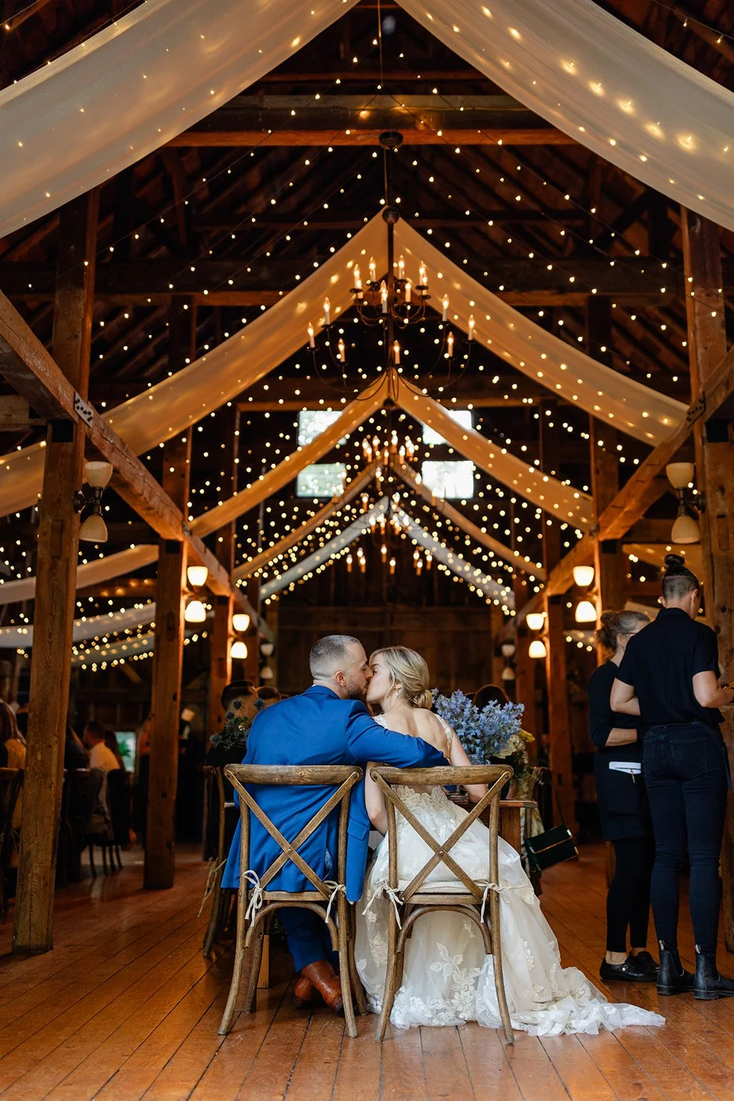 A bride and groom sharing a kiss at their wedding reception in a rustic barn decorated with string lights and draped fabric.