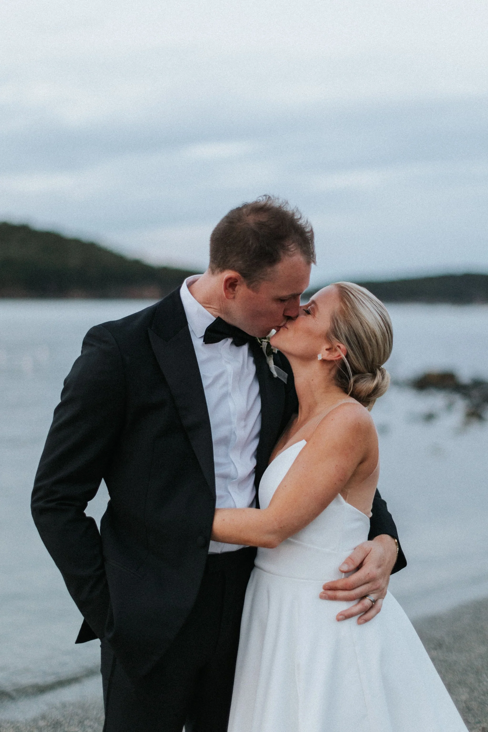 A newlywed couple sharing a kiss by the water on their wedding day, with calm water and distant hills in the background.