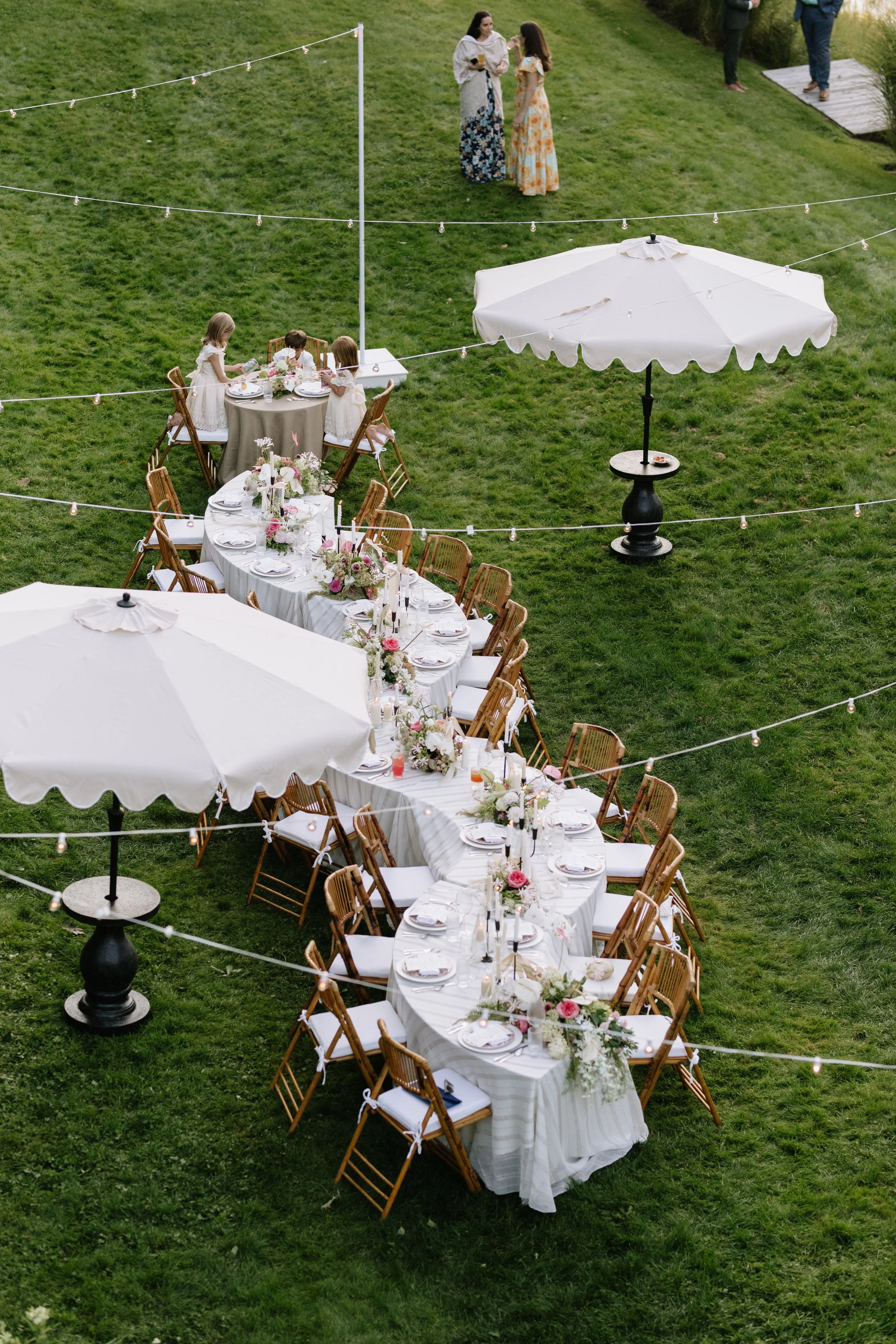 A long outdoor dining table set with white tablecloths, floral centerpieces, and elegant place settings, located on a grassy lawn with large white umbrellas and string lights overhead. In the background, children in white dresses sit at a smaller tab