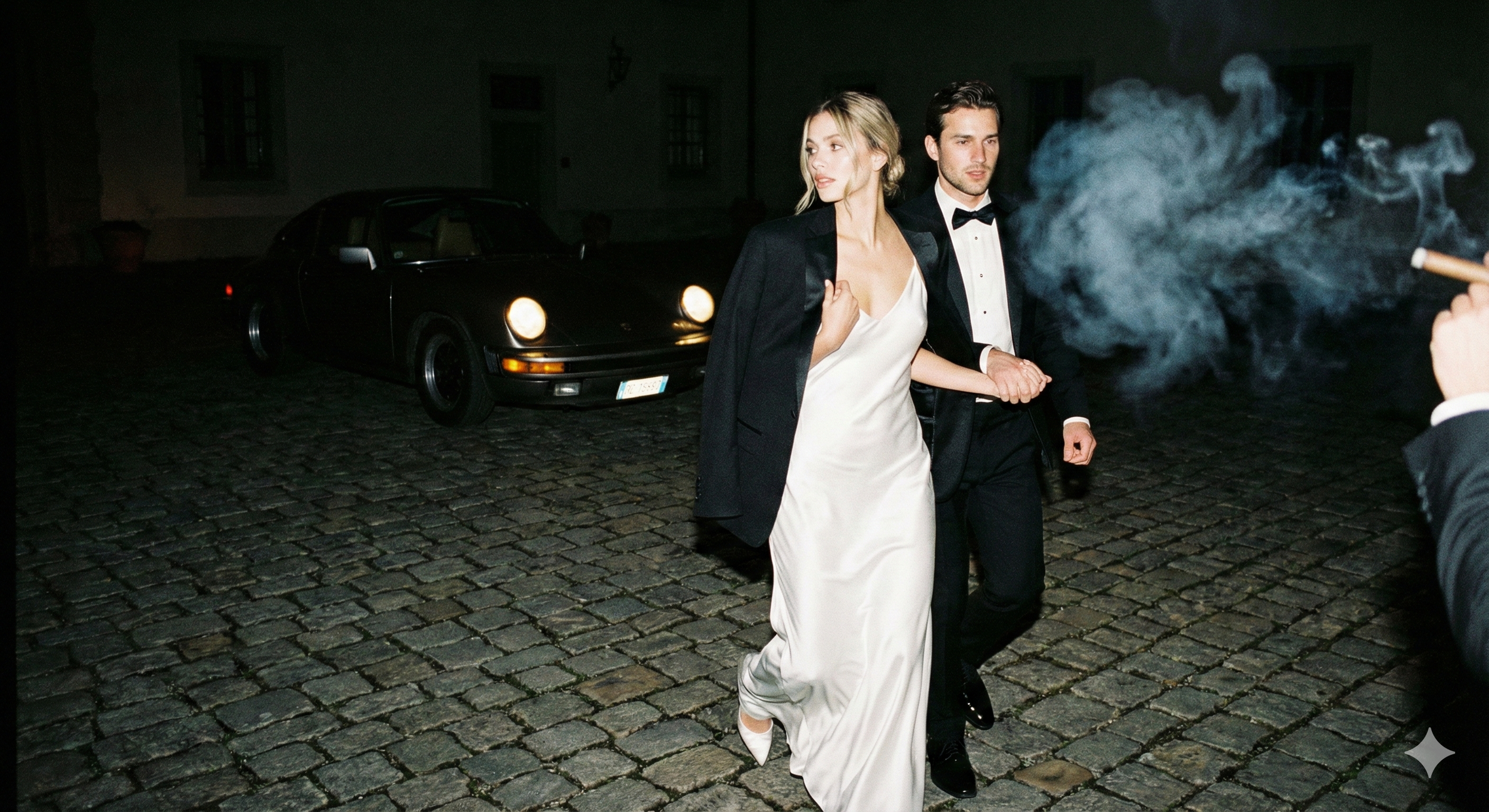 A man and woman in formal attire walking on cobblestone street at night, with a black vintage car in the background and someone smoking a cigarette nearby.