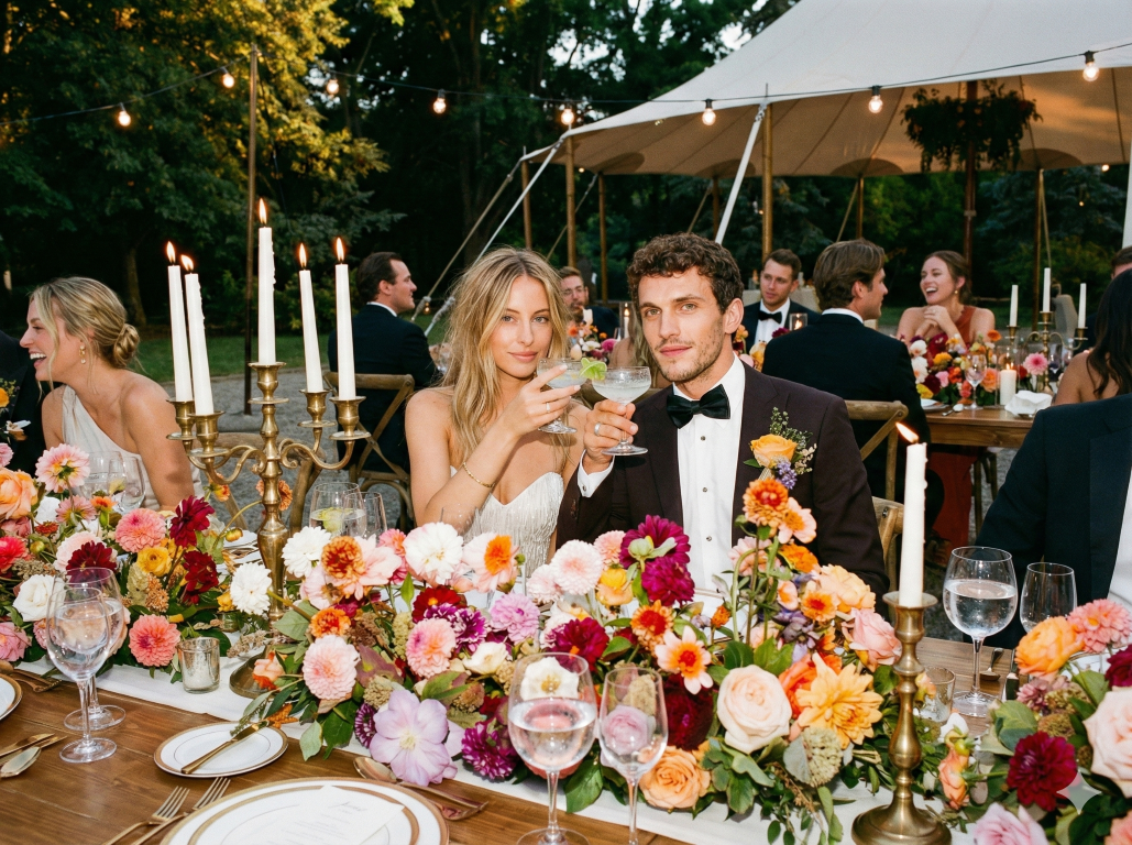 A wedding reception with a bride and groom toasting, surrounded by floral arrangements, candles, and guests in formal attire under string lights and a tent.