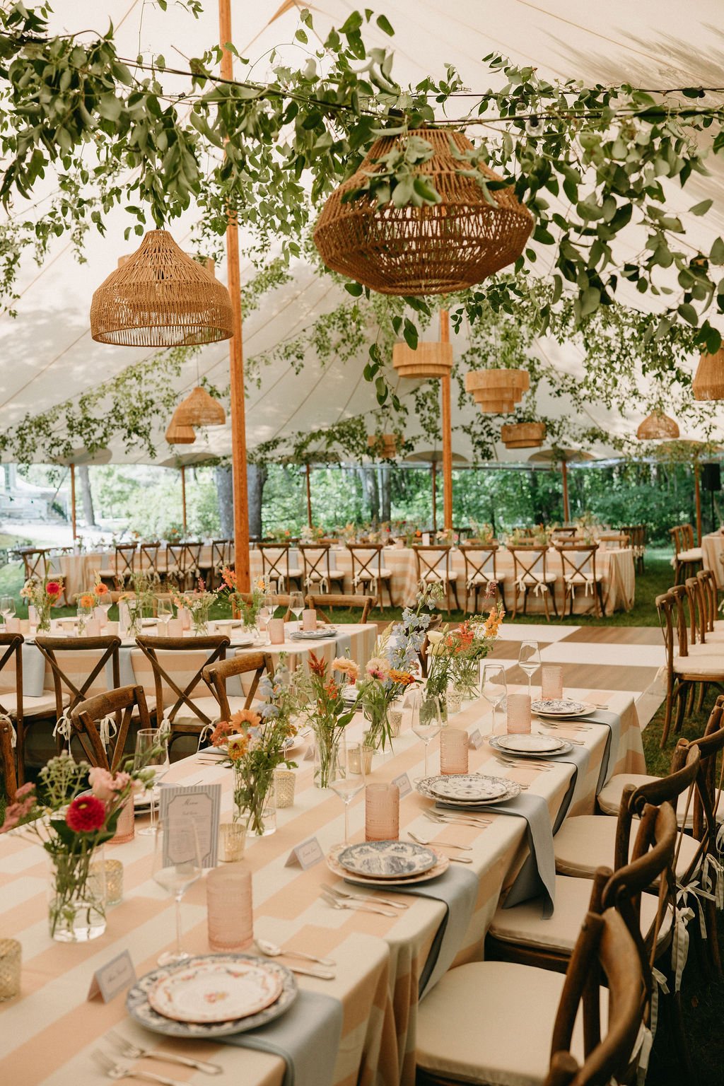 Decorated outdoor dining area under a large tent with hanging wicker light fixtures and lush greenery, featuring long tables with floral centerpieces and individual place settings.