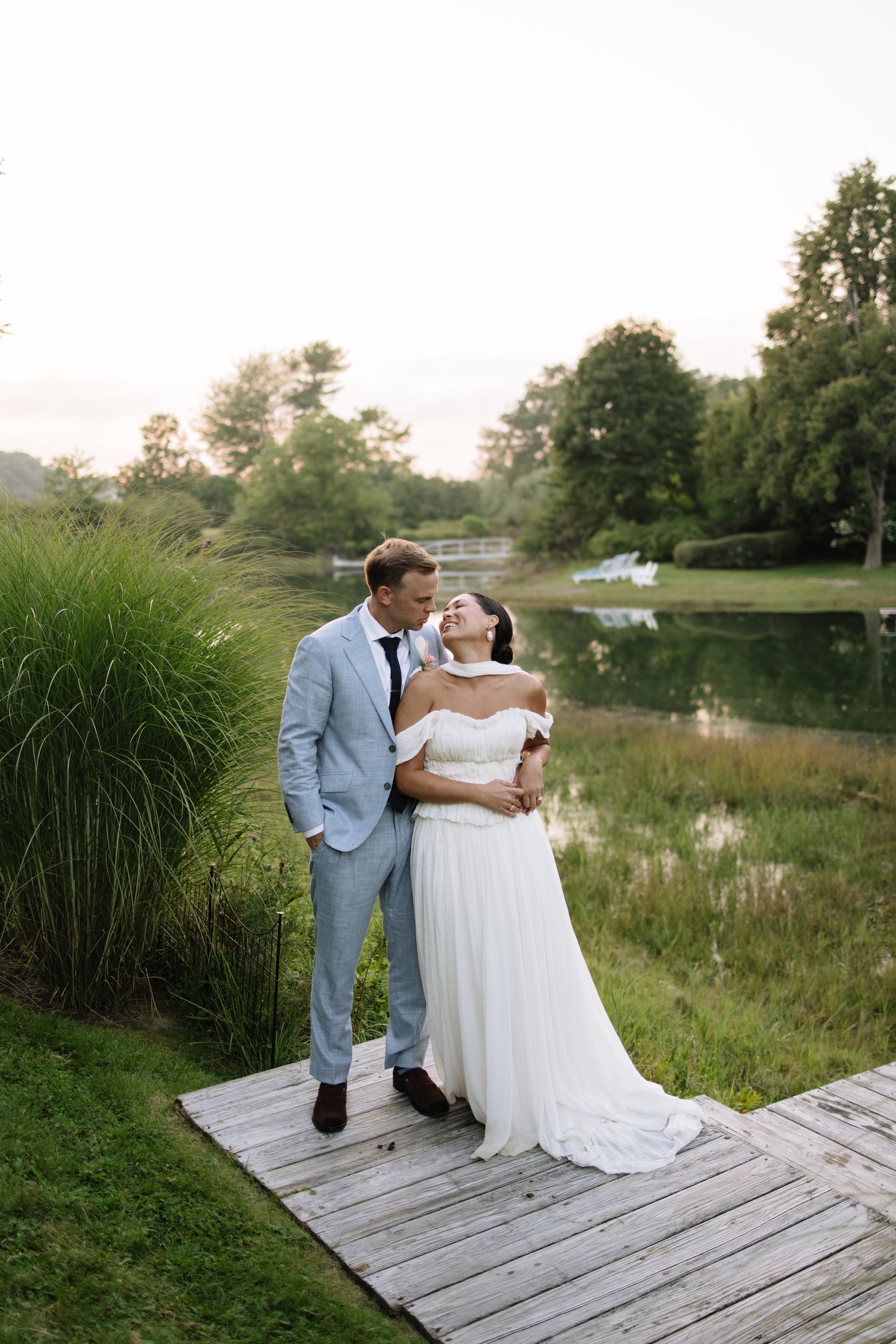 A bride and groom standing on a wooden dock by a lake, smiling at each other, with trees and a grassy landscape in the background during daytime.
