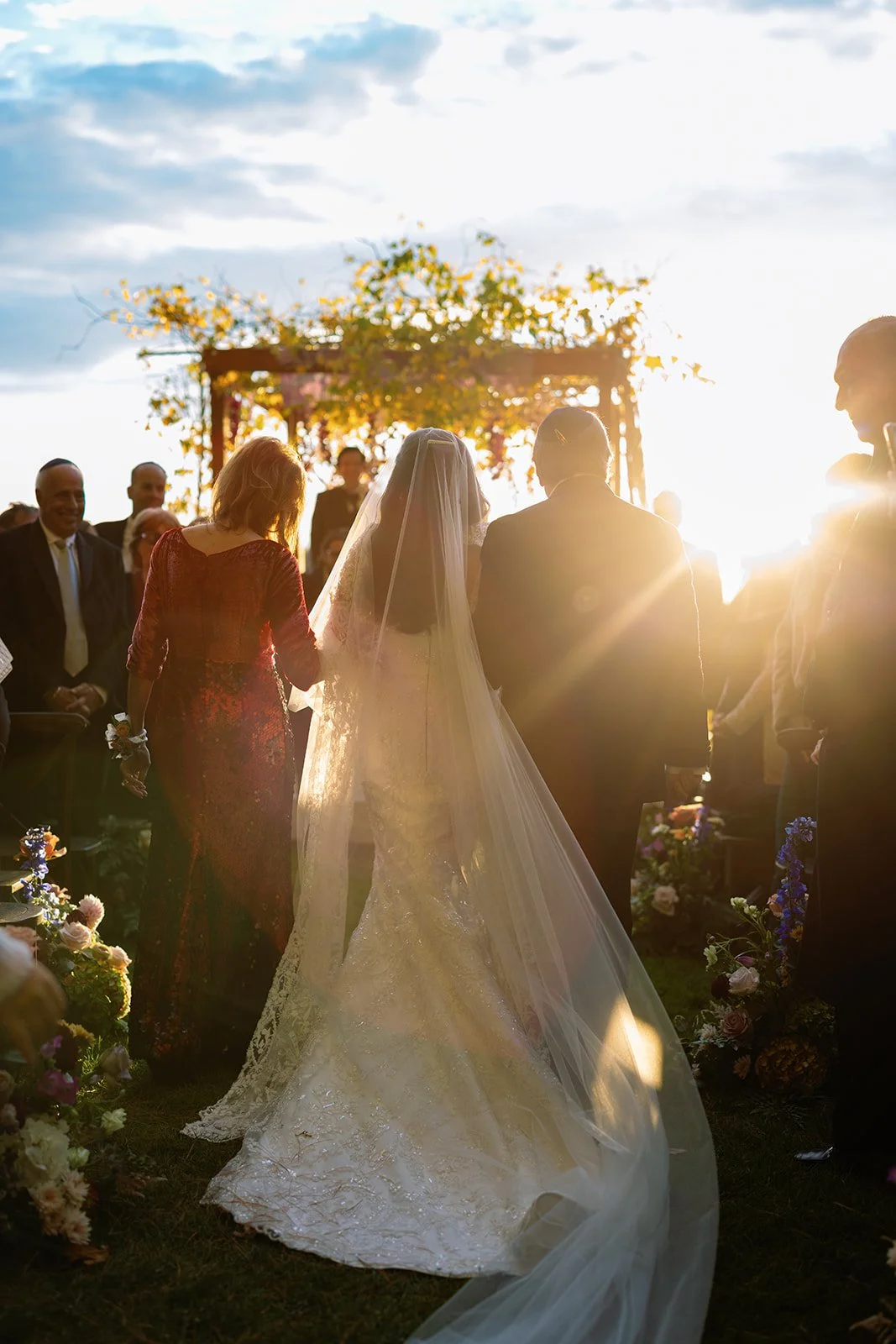 Bride walking down the aisle with her parents during an outdoor wedding at sunset.