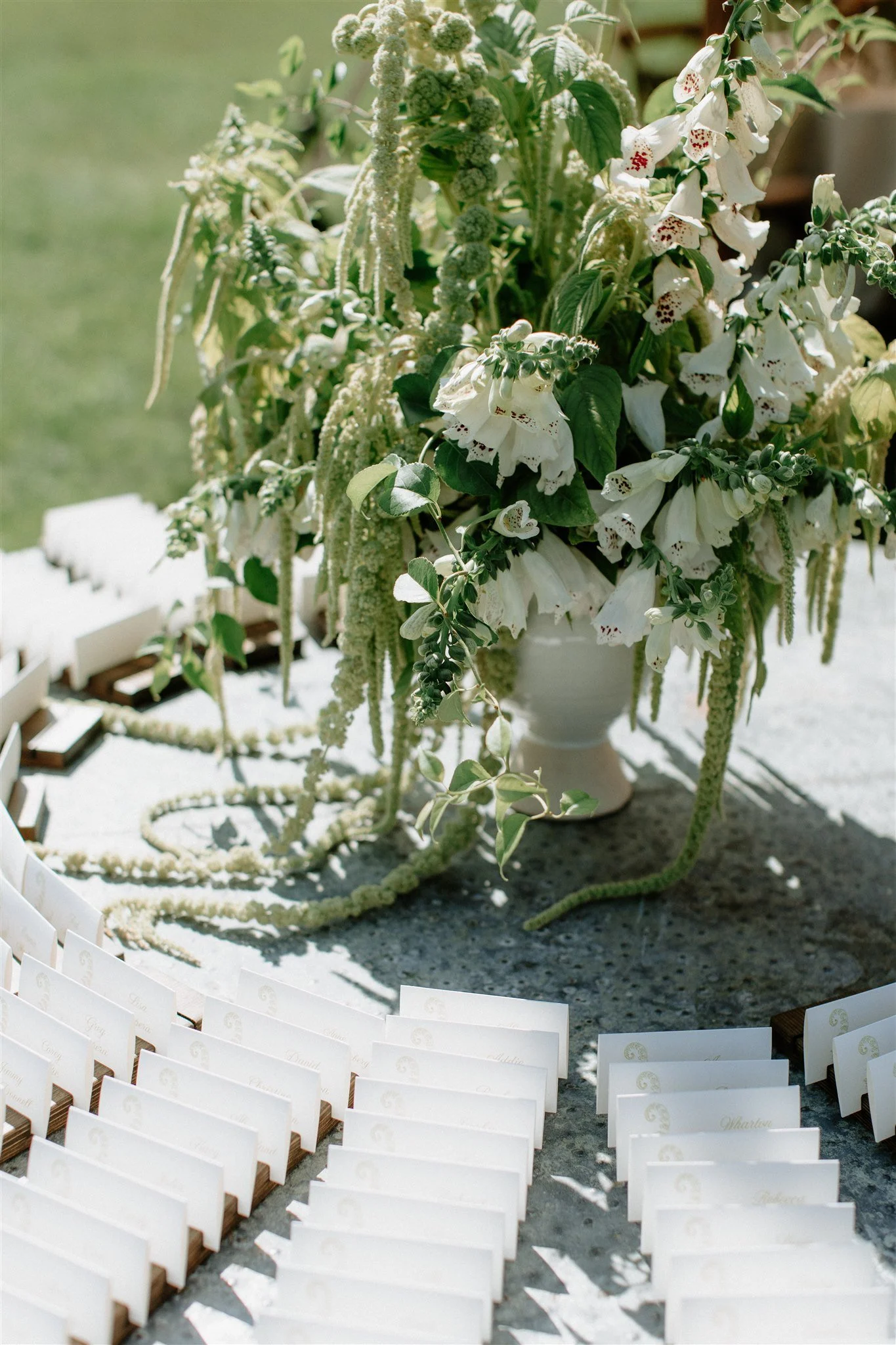 A floral arrangement with white flowers in a white vase on a table, surrounded by small, white, seat cards with names on a gravel surface outdoors.
